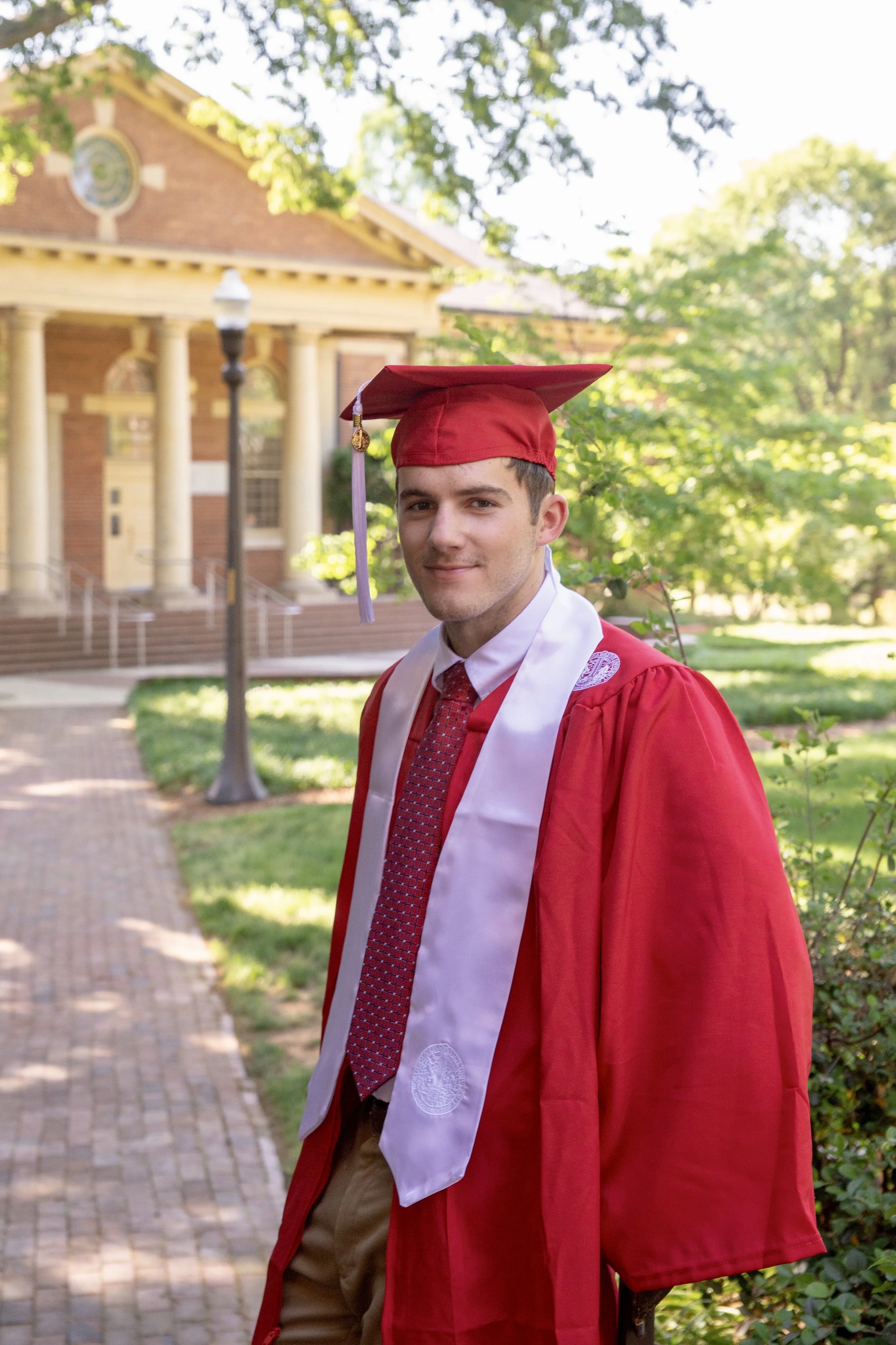 A young man in a red graduation gown and cap with a white stole, standing outside on a brick pathway in front of a college campus building, smiling at the camera during daylight.