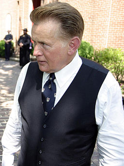 Actor Martin Sheen, playing Josiah Bartlet, is standing on the set of The West Wing. He is outside of St. Anne's Church. There is a row of greenery behind him.