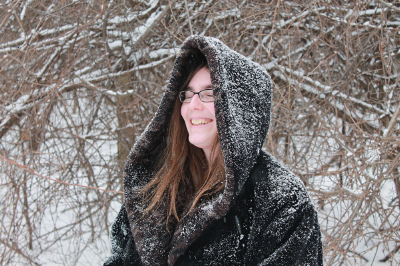 Ada Hoffmann standing in a winter forest. The trees behind her have snow, with brown bark peaking through. She is wearing a heavy black coat with the hood pulled up over her head. Her honey brown hair can be seen out of the front of her hood, framing a smiling face.