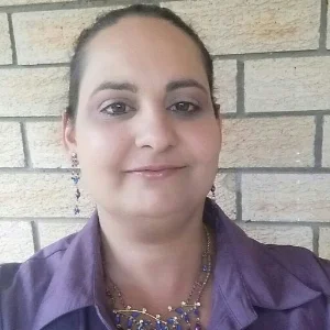 Karletta Abianac, dark hair pulled back, wearing long dangling earrings, is standing in front of a beige brick wall. She is wearing a purple blouse, and matching nechlace.