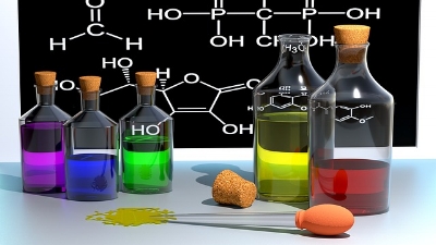 Chemistry lab table, with 3 small bottled and 2 large ones full of colorful liquid. On a chalkboard behind them are various chemical formulas.