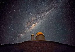 The ESO telescope located at La Silla, Chile. The building has a gold dome, and is sitting on a hill. In the background is the milky way.