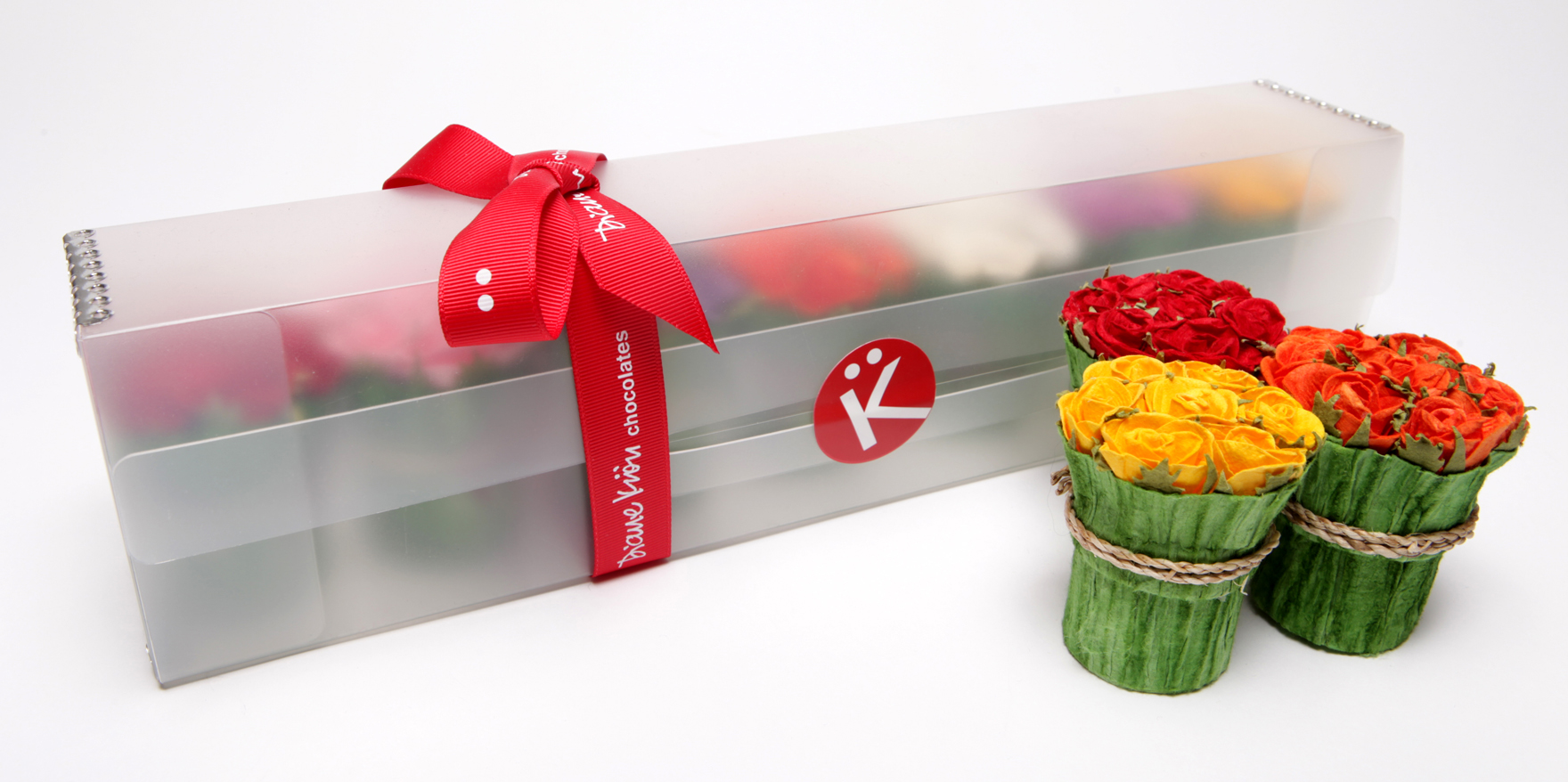  Clear Box tied with red ribbon holding seven mini bouquet boxes of chocolate in background. Foreground shows three mini bouquet boxes with red yellow and orange flowers 