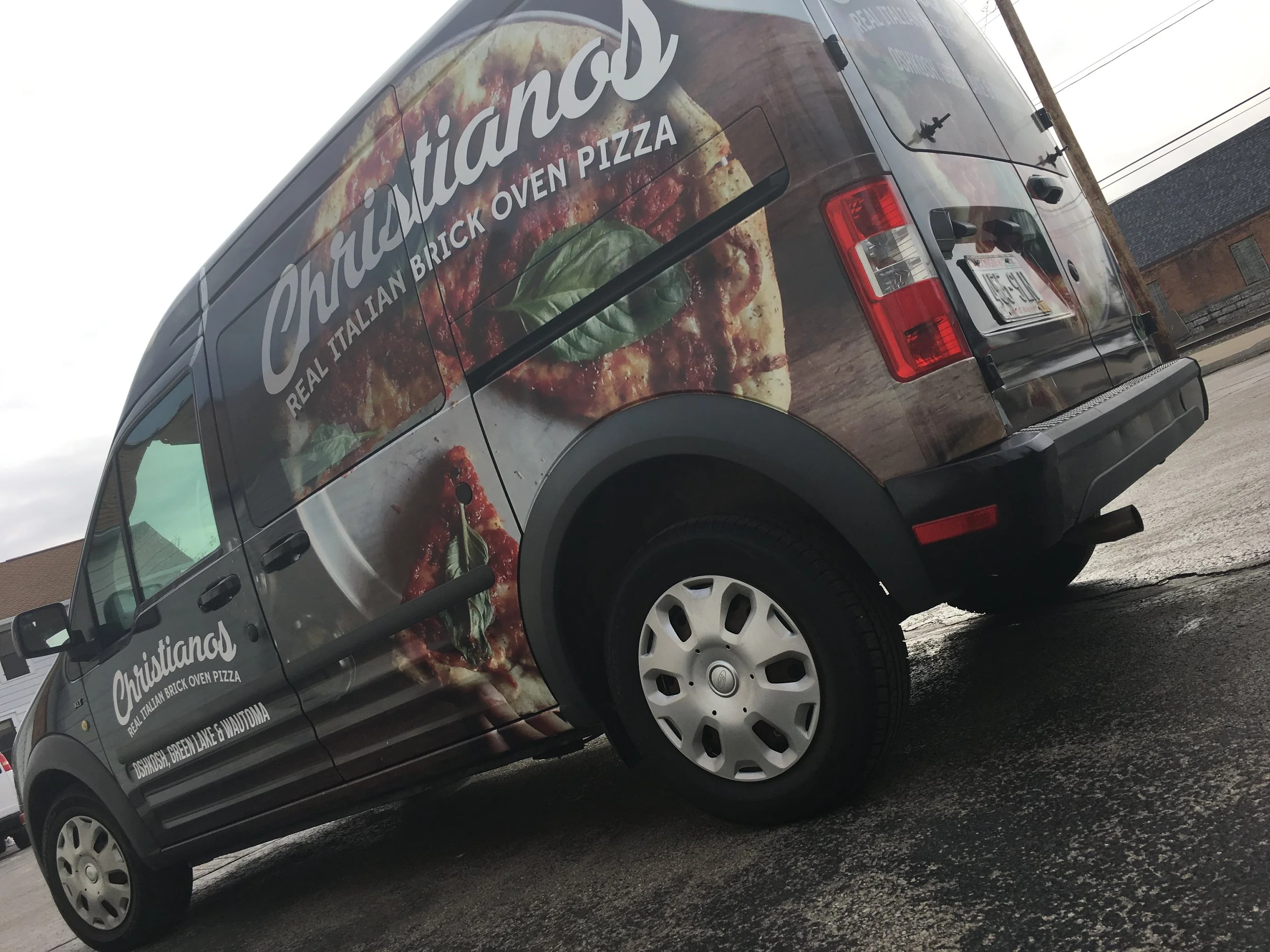A black pizza delivery van with large images of pizza and basil leaves on its side, advertising Christiano's Italian brick oven pizza.