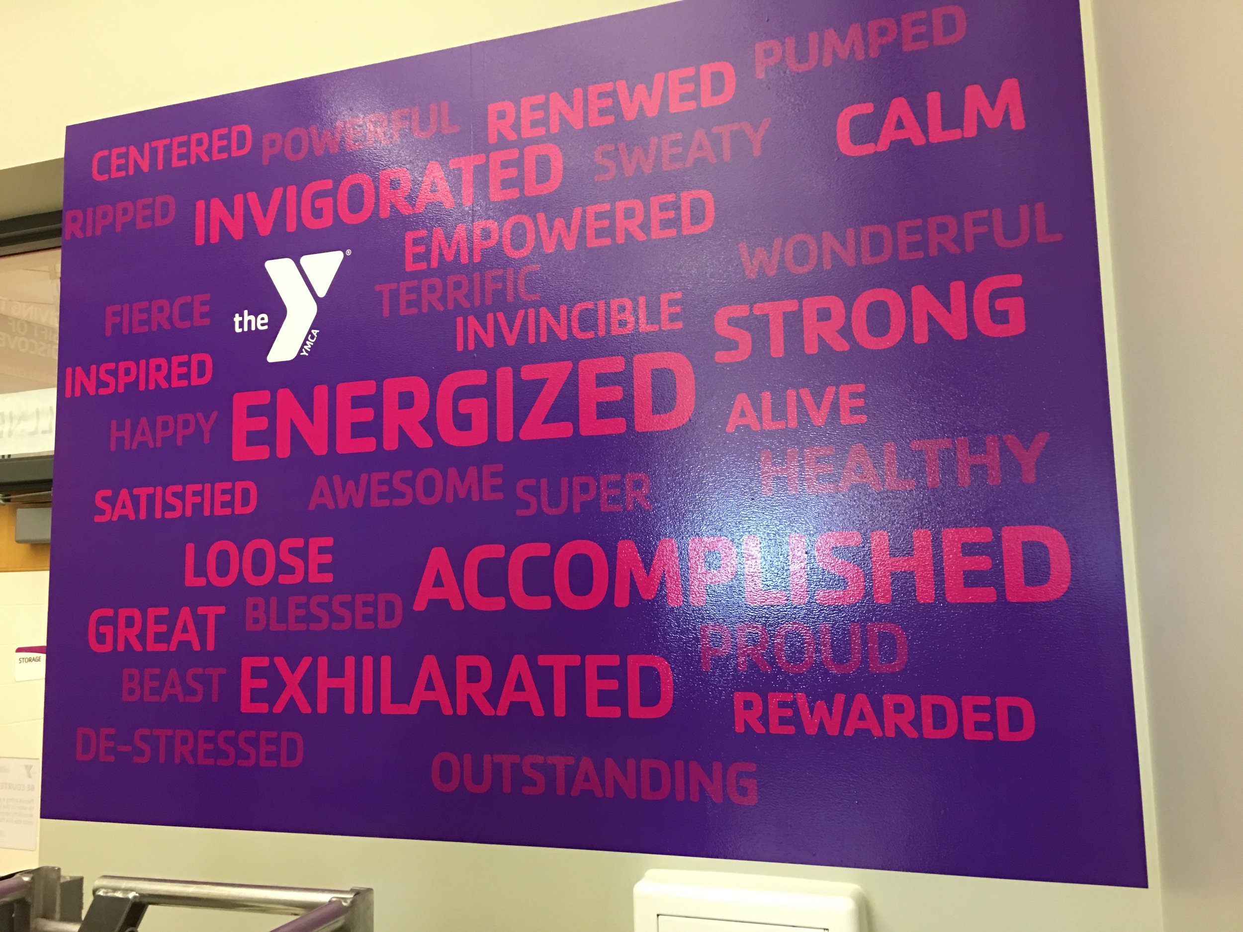 A purple wall displaying positive words associated with the YMCA, including 'ENERGIZED,' 'proud,' 'accomplished,' 'strong,' 'healthy,' and 'outstanding,' with a white YMCA logo near the top left.