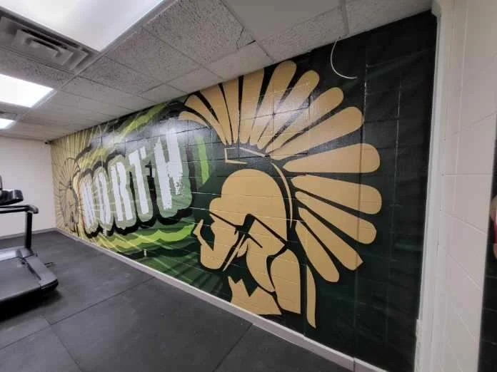 A wall mural in a gym featuring a stylized profile of a person with a mohawk hairstyle, wearing a cap, with sunburst rays behind their head, and the word 'DROPP' in bold letters to the left.