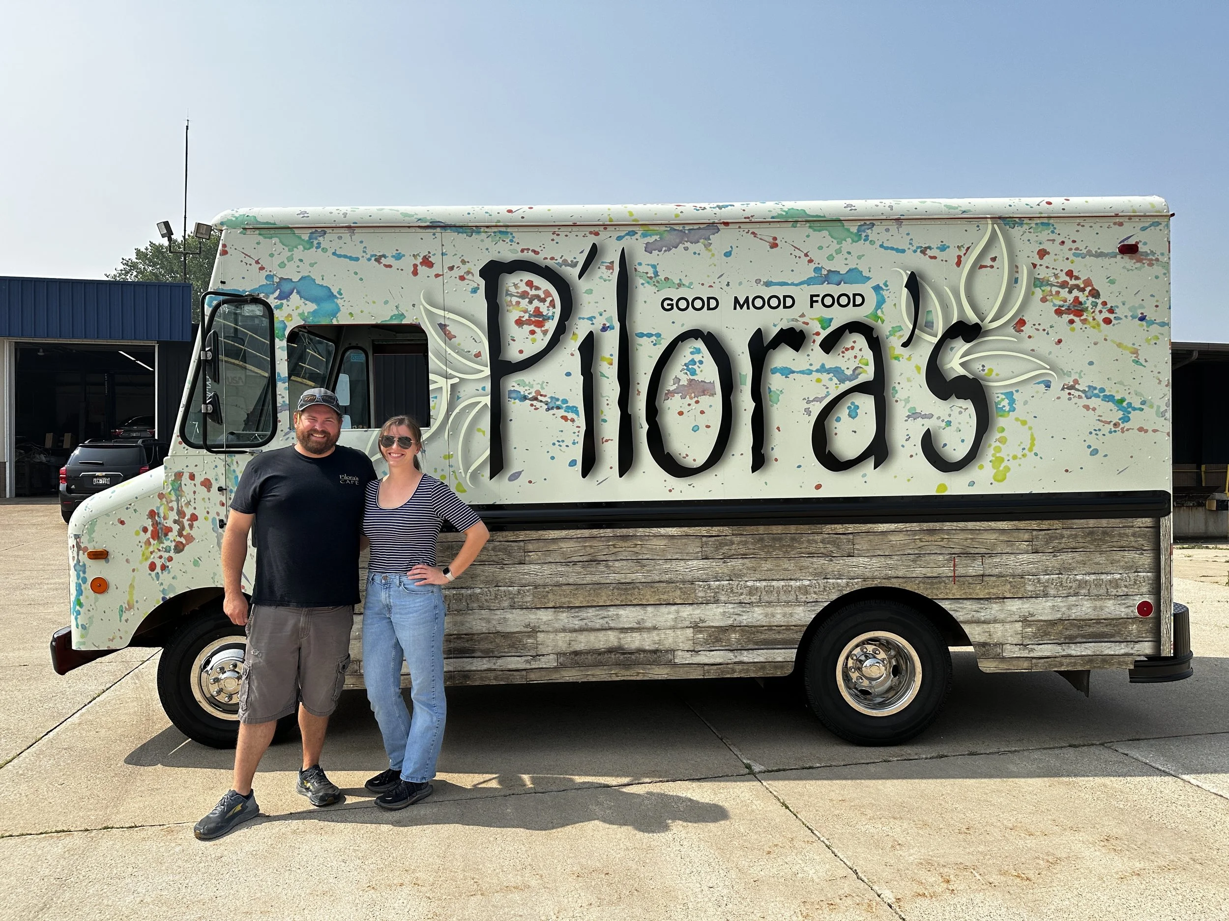 Two people, a man and a woman, smiling and standing in front of a colorful food truck named 'Pioras' with the words 'Good Mood Food' written on it. The truck has a paint splatter design on the top and a wood panel section at the bottom.