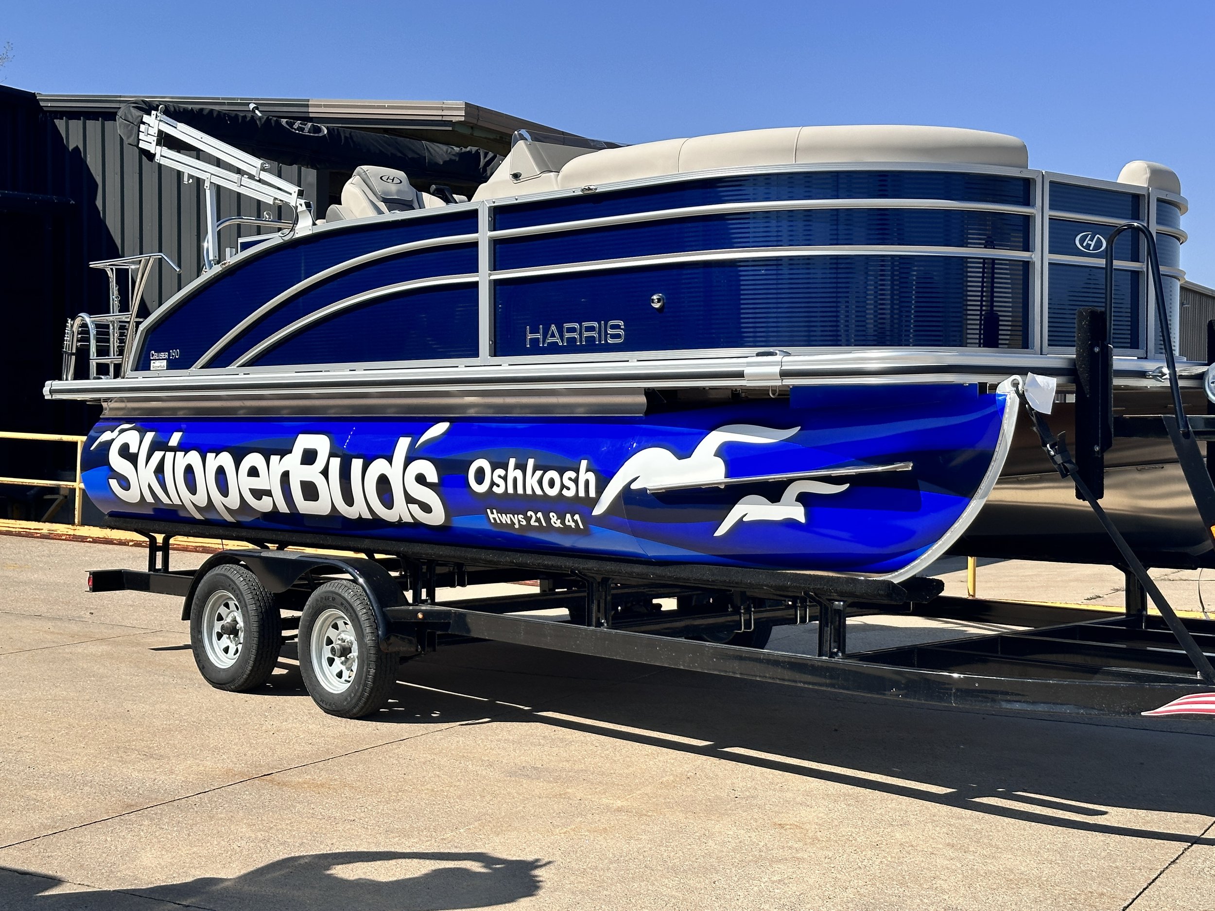 A blue and gray pontoon boat named 'Skipper Buds' parked on a trailer in an outdoor lot, with a building and a clear blue sky in the background.