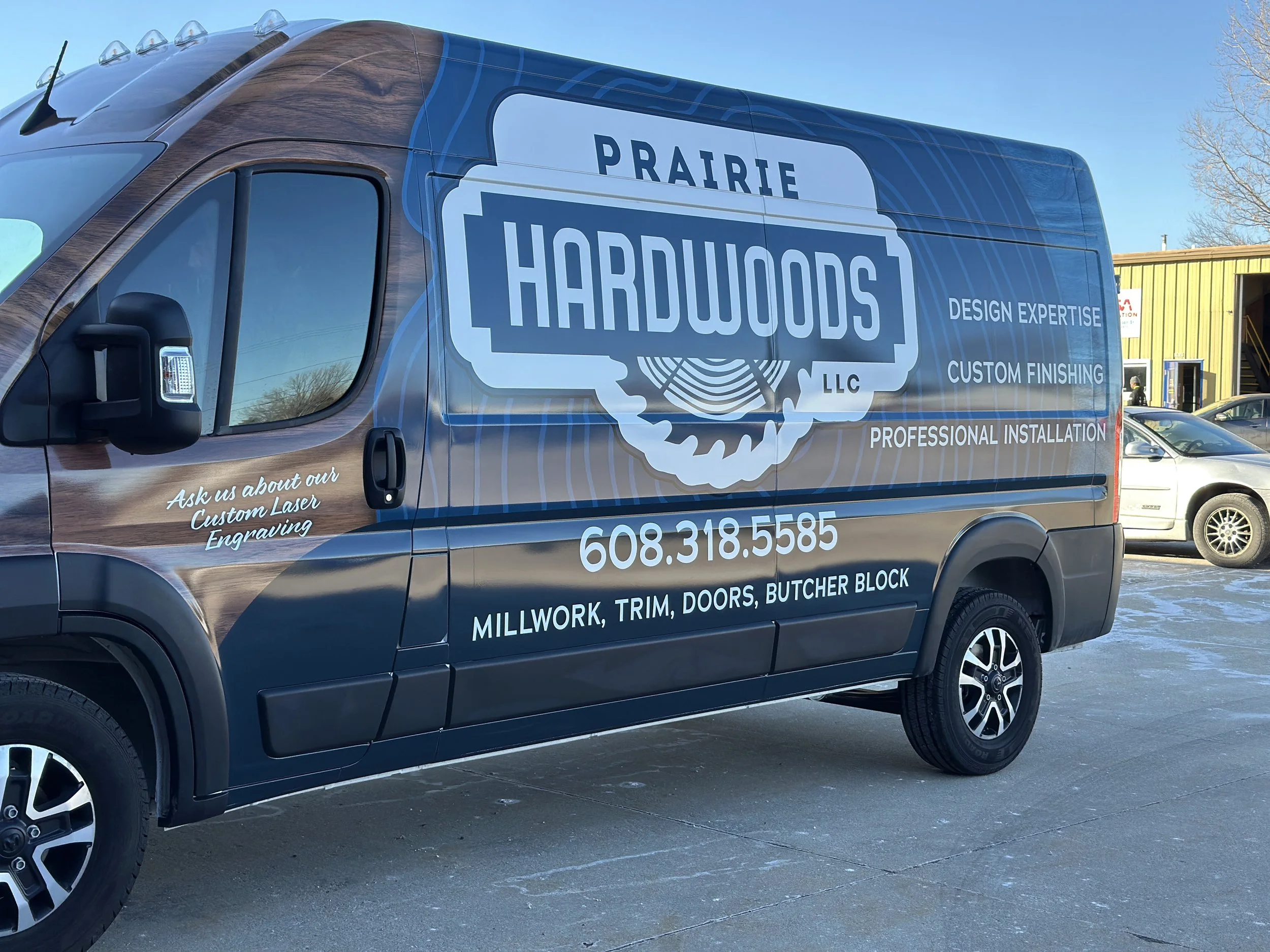 A commercial van with a wood grain design on part of the side, advertising Prairie Hardwoods LLC. It lists services including design expertise, custom finishing, and professional installation. The van displays the phone number 608-318-5585 and mentions millwork, trim, doors, and butcher block. There is also a message about custom laser engraving.