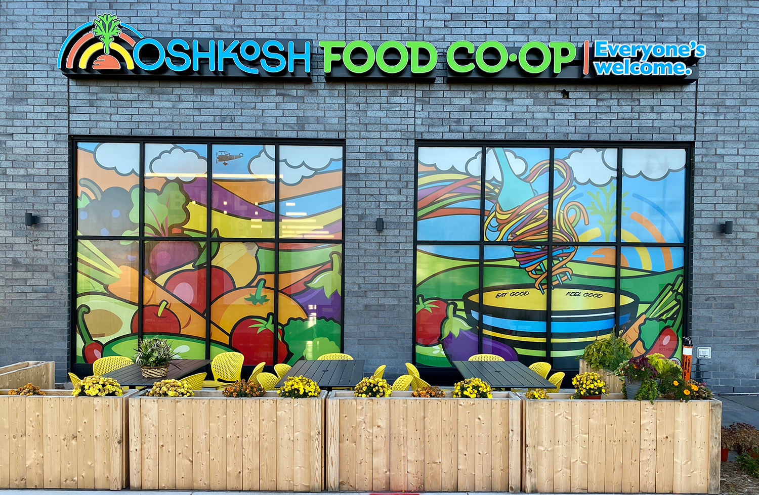 Exterior of OSHKOSH FOOD CO-OP store with colorful mural windows featuring vegetables and food items, yellow chairs, and wooden planters with flowers in front.