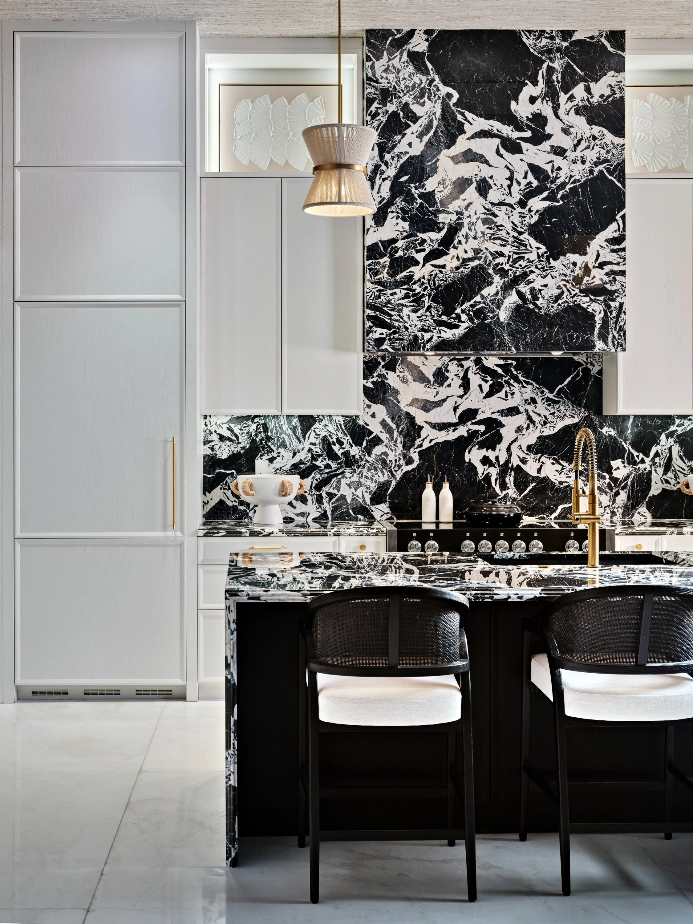 Modern kitchen with black and white marble backsplash and countertops, white cabinets, black bar stools with white cushions, and a gold faucet.