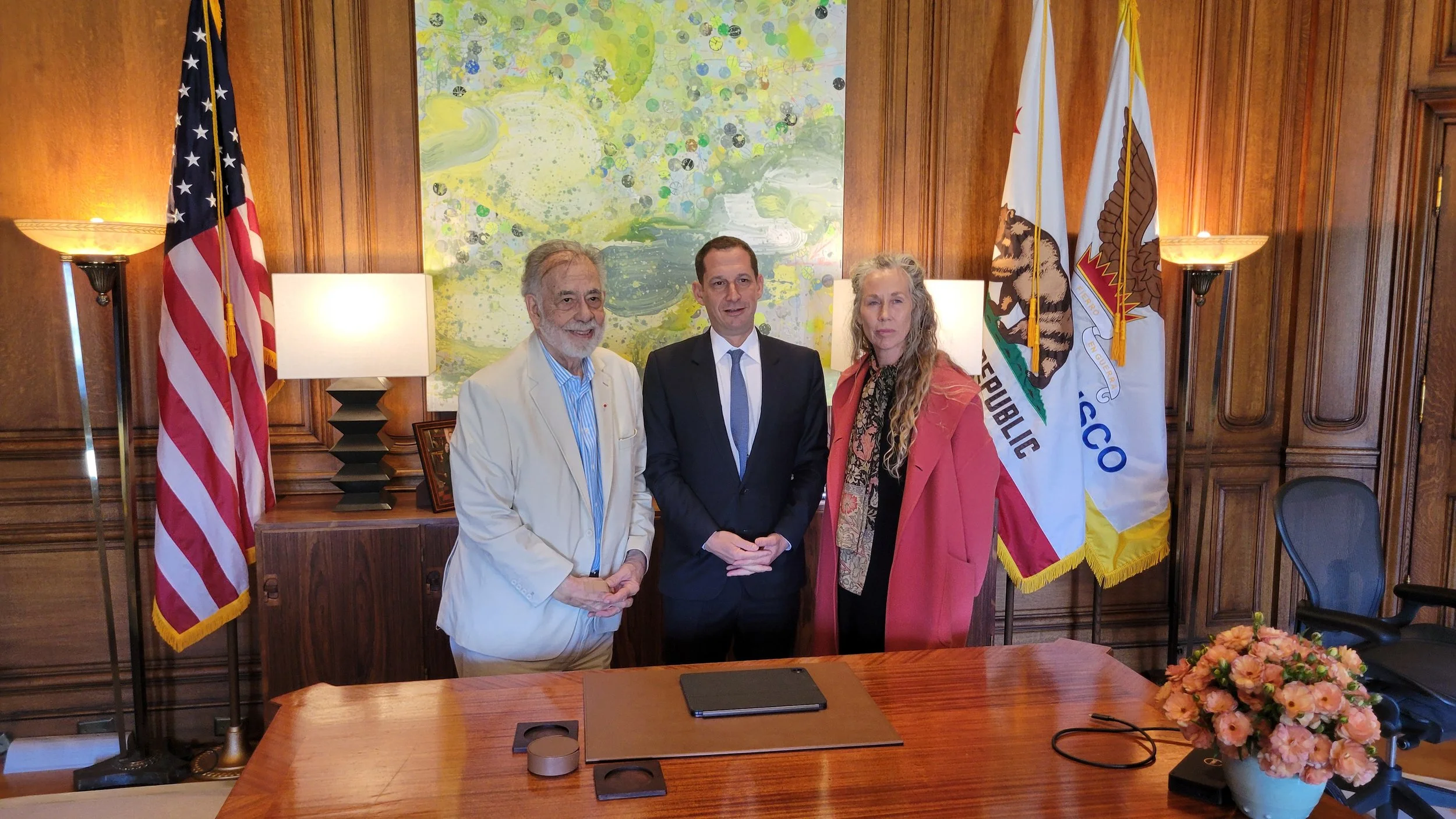 Three individuals standing in a wood-paneled room with American and California flags and a colorful abstract painting in the background.