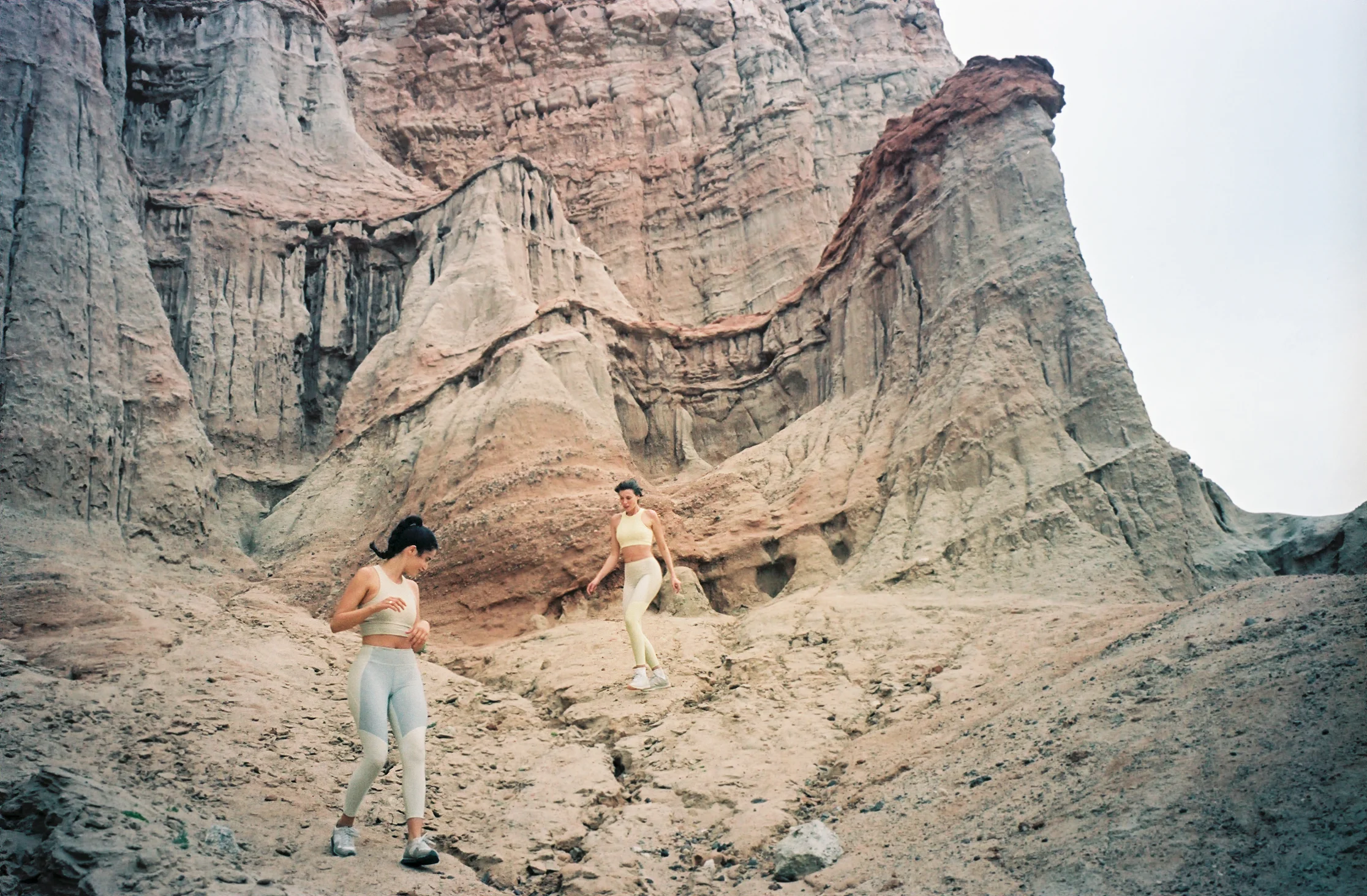 Two women in athletic wear hiking in a rocky, mountainous landscape with steep cliffs and a cloudy sky.
