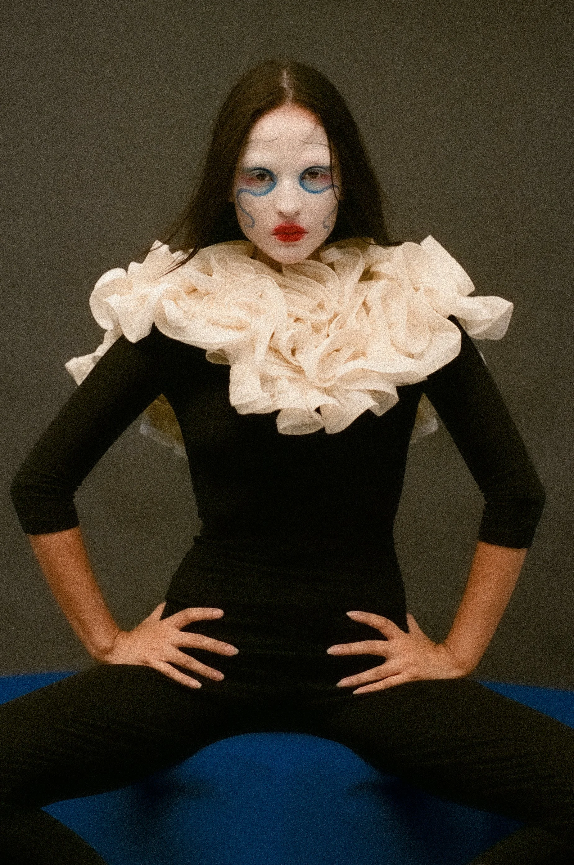 A woman in a black dress with a large, ruffled, cream-colored collar poses with hands on her hips against a plain dark background.
