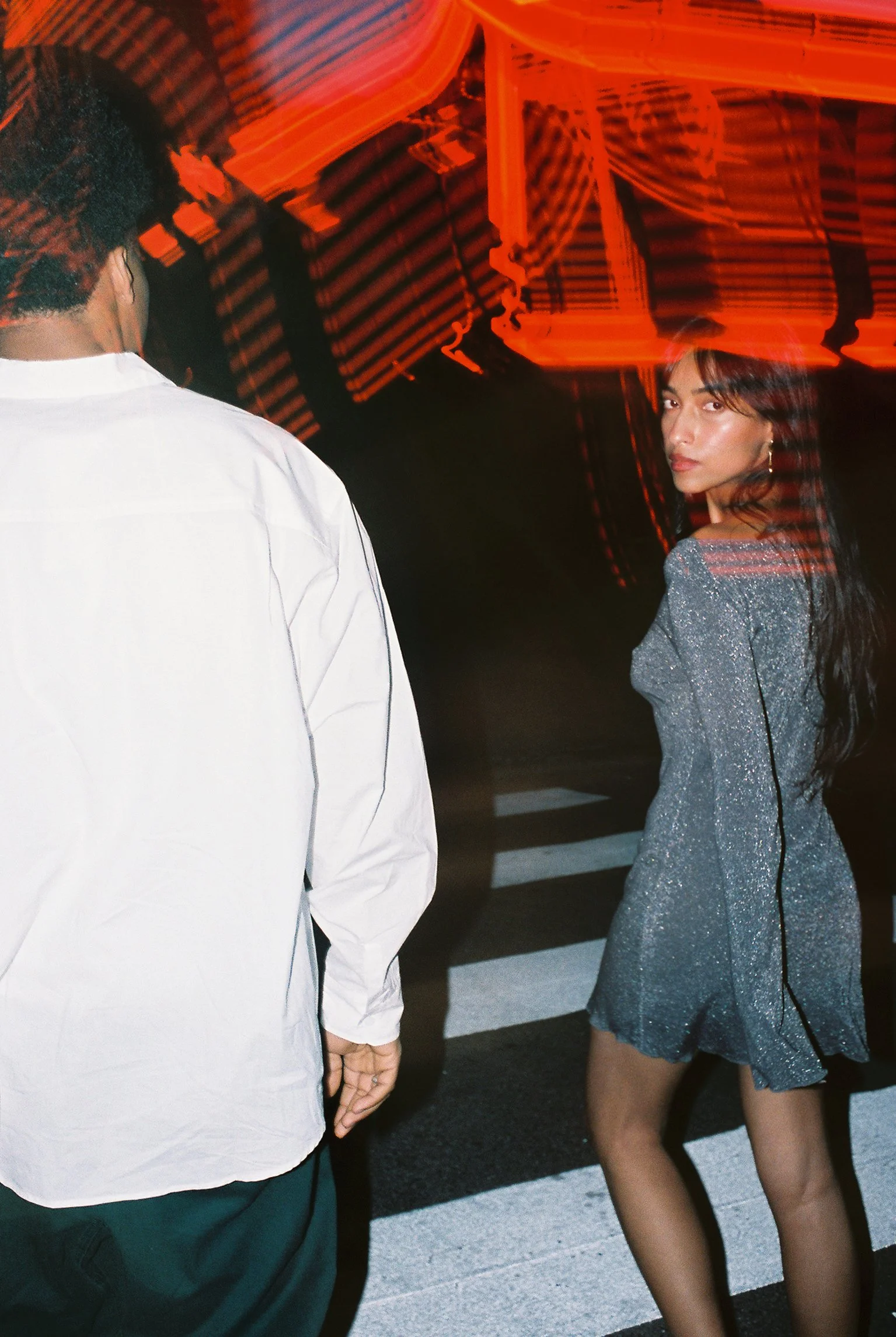 A woman with dark hair and light skin dressed in a shimmery, short, off-the-shoulder dress stands on a crosswalk at night, looking back at the camera with a serious expression. A person with short curly hair wearing a white shirt is walking away from