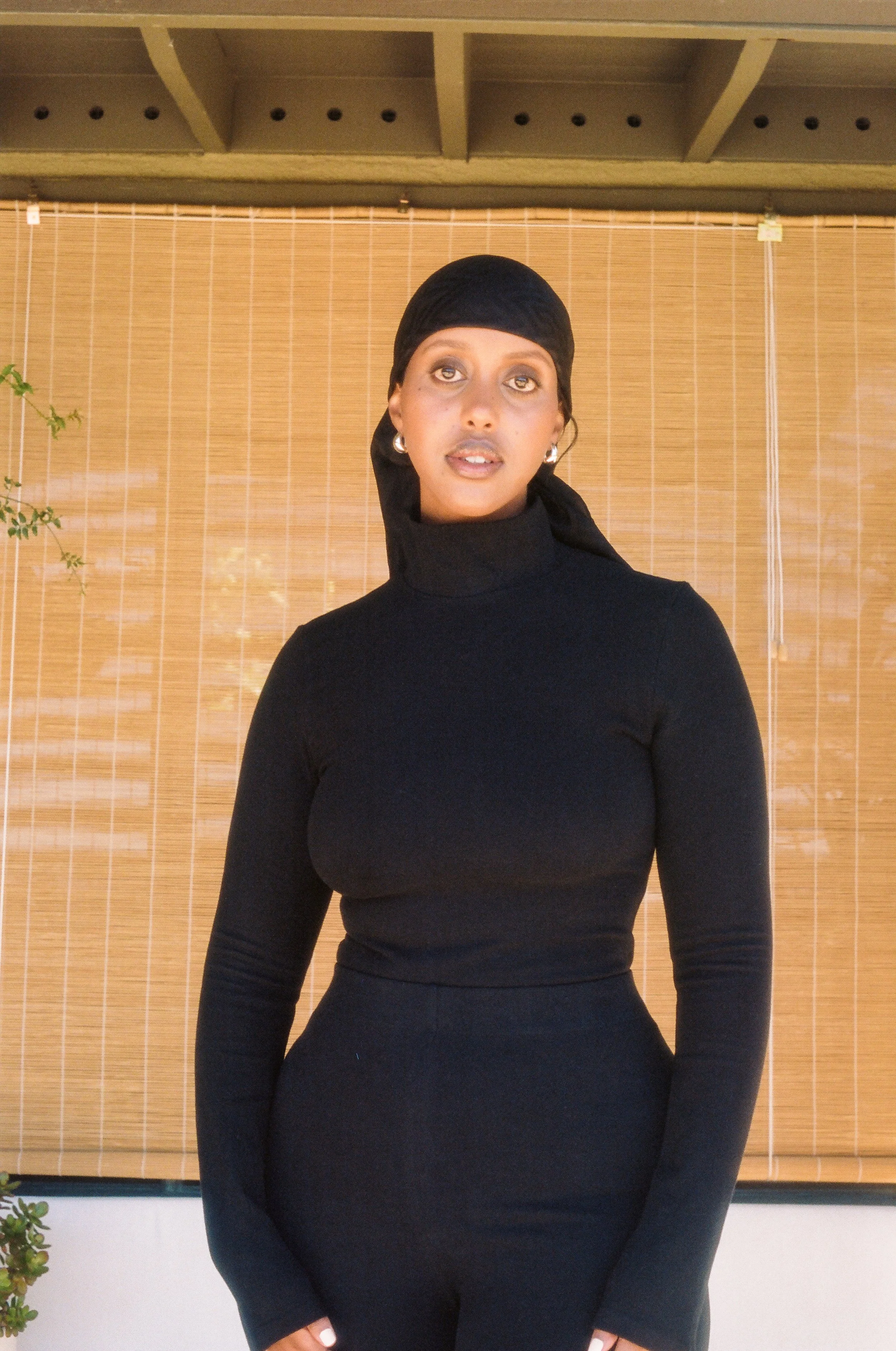 A woman with dark hair, wearing a black turtleneck and earrings, standing in front of a bamboo shade.
