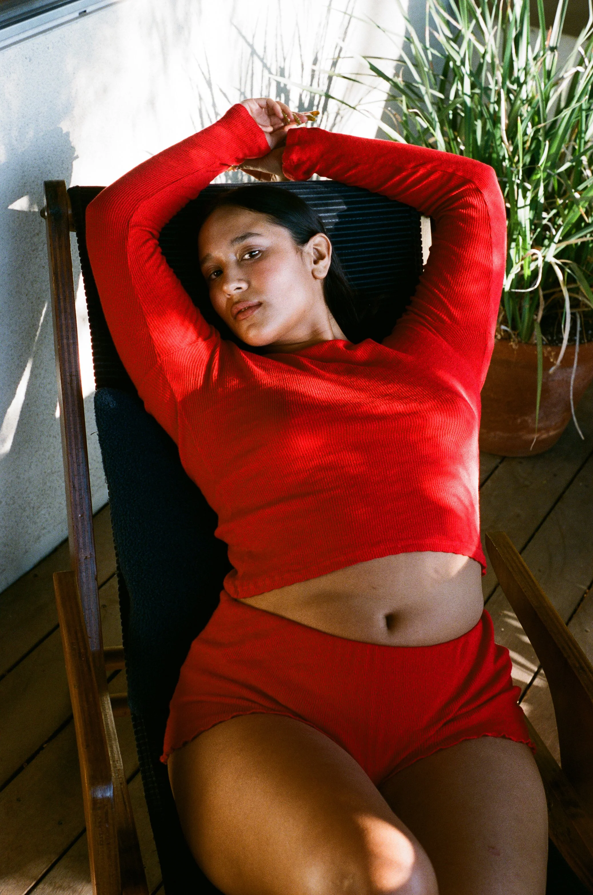 A woman in red lounge wear lying on a black outdoor lounge chair, with a potted plant in the background.