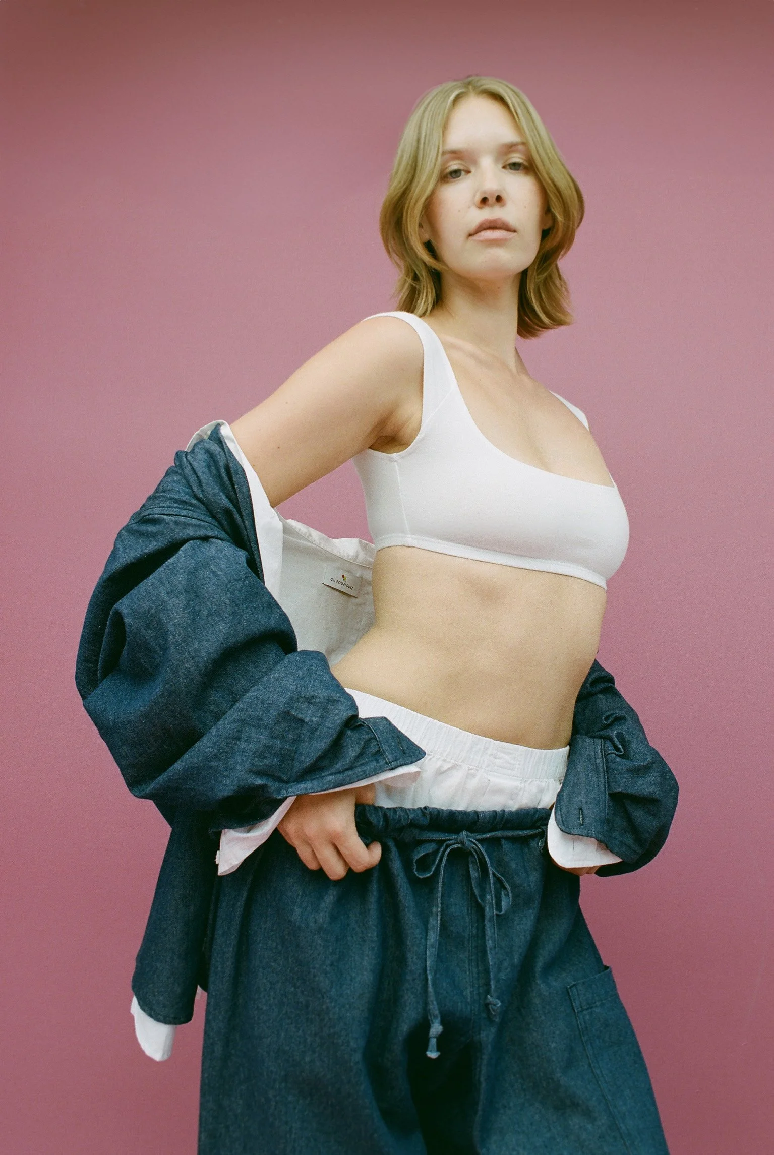 Woman with shoulder-length blonde hair standing against a pink background, wearing a white crop top and dark loose-fitting pants, with a denim jacket slipping off her shoulders.