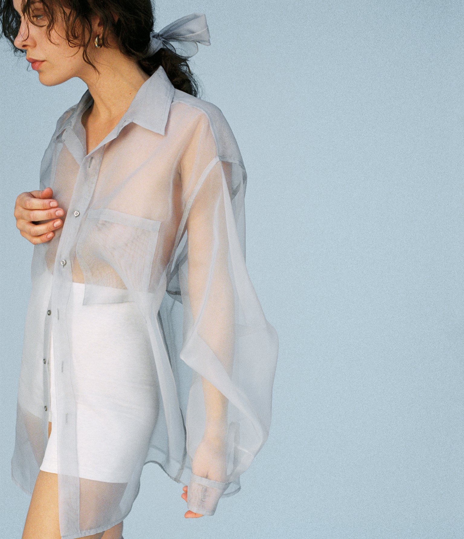 Woman wearing a sheer button-up shirt and white shorts, with a gray bow in her hair, standing against a light blue background.