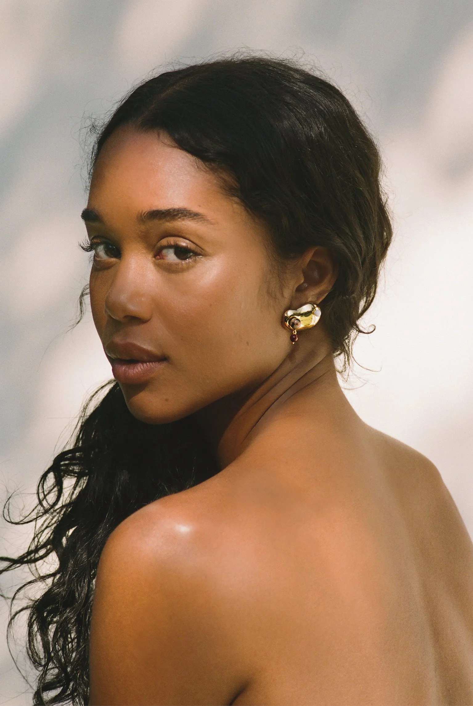 A woman with dark, wavy hair looking over her shoulder at the camera, wearing gold earrings and minimal makeup, with her bare shoulder visible.