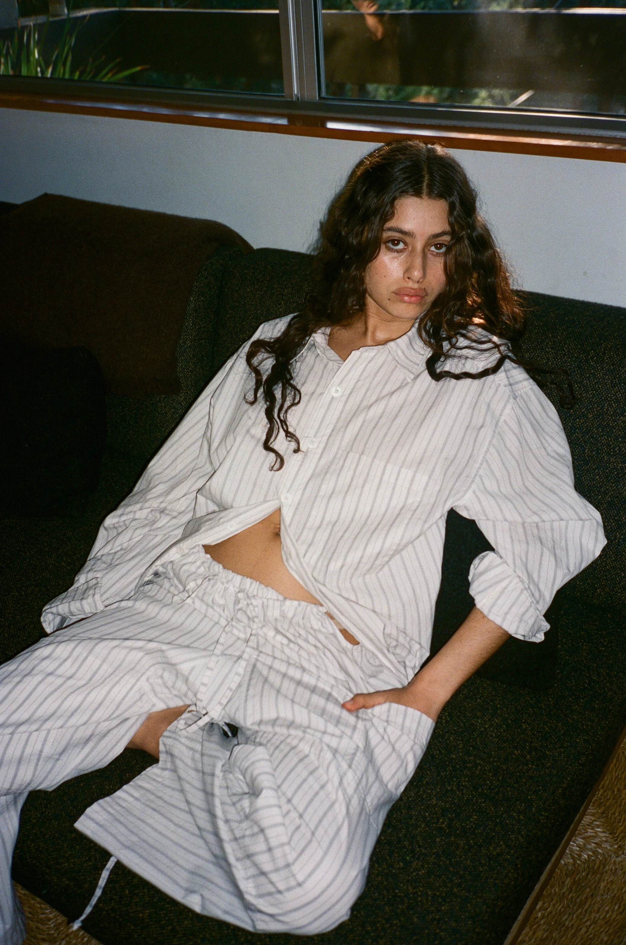 A young woman with long curly hair and light skin, wearing striped pajamas, is sitting on a dark-colored couch in a room with a window in the background.