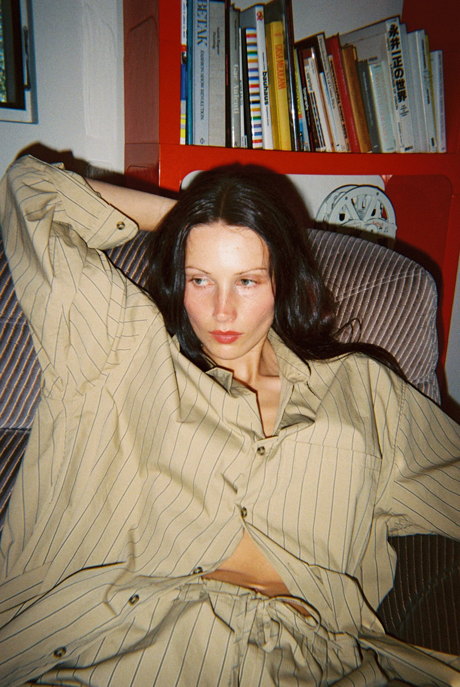 A woman with long dark hair lying on a striped bed, wearing a beige striped shirt and matching pants, with bookshelves in the background.