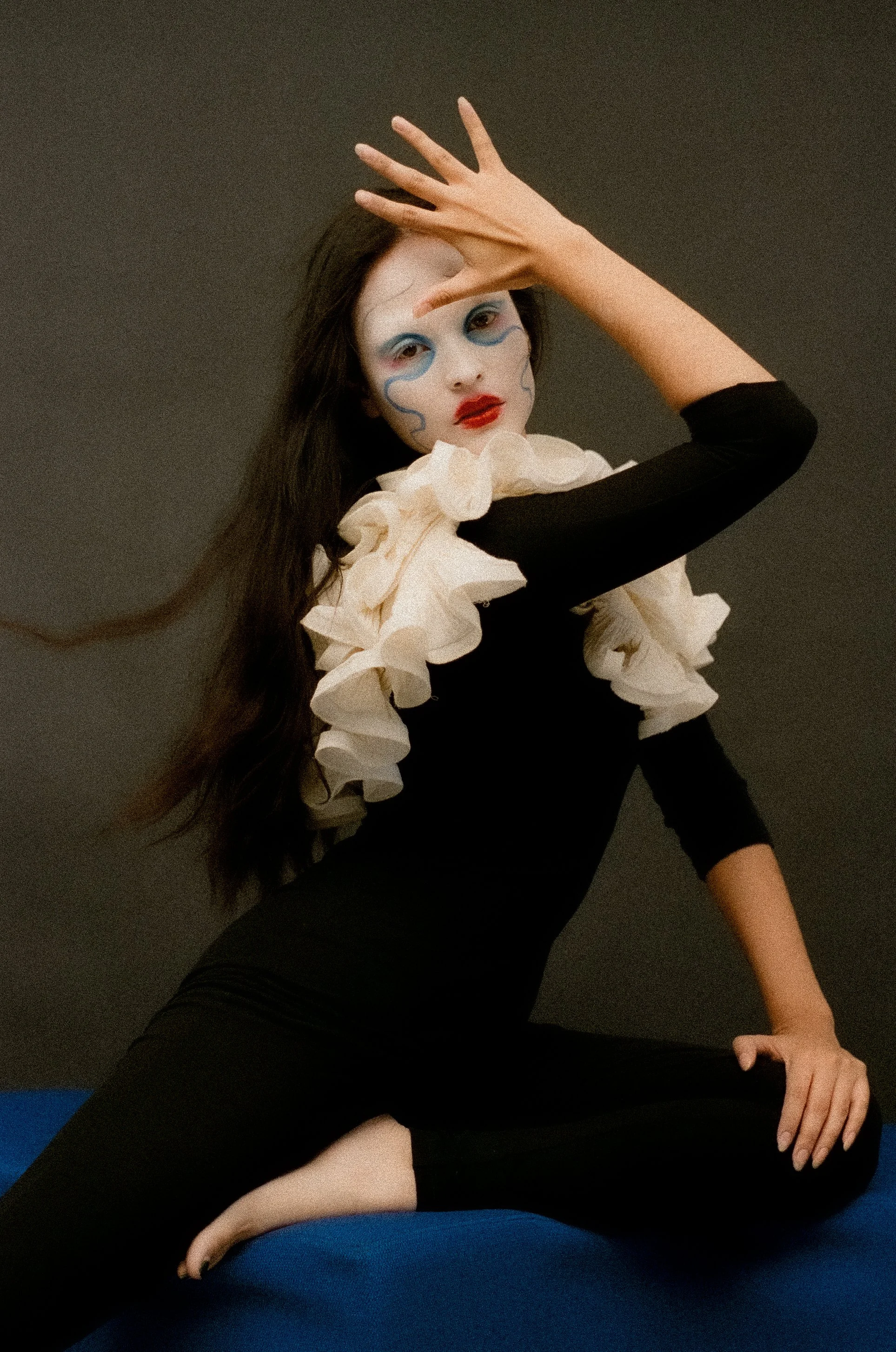 A woman with dramatic makeup, long dark hair, and a white ruffled collar, dressed in black, posing against a dark background with one hand on her forehead and the other on her knee.