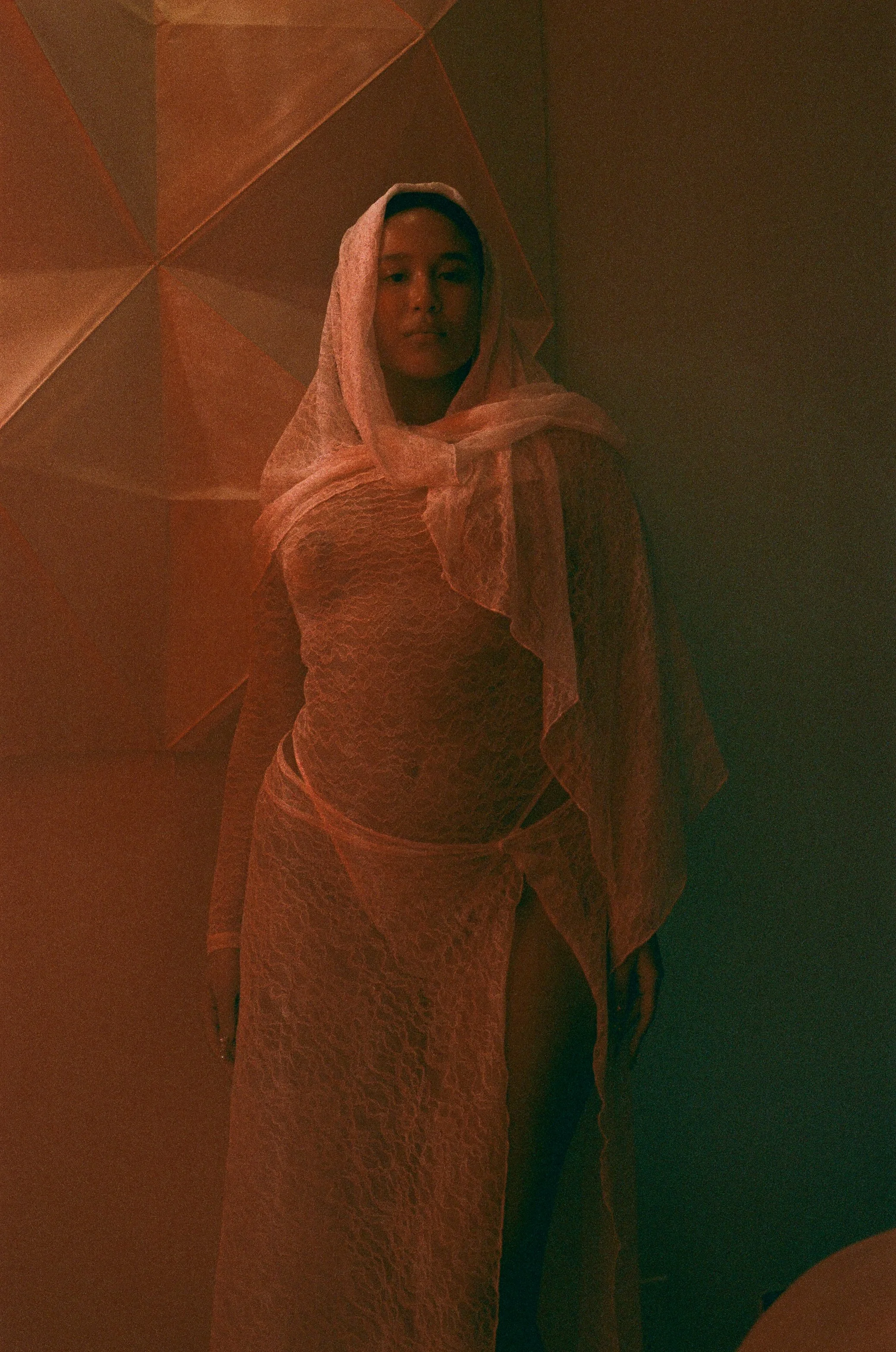 A person standing against a background of geometric paneling, draped in sheer lace fabric with a matching headscarf, illuminated by warm, reddish lighting.