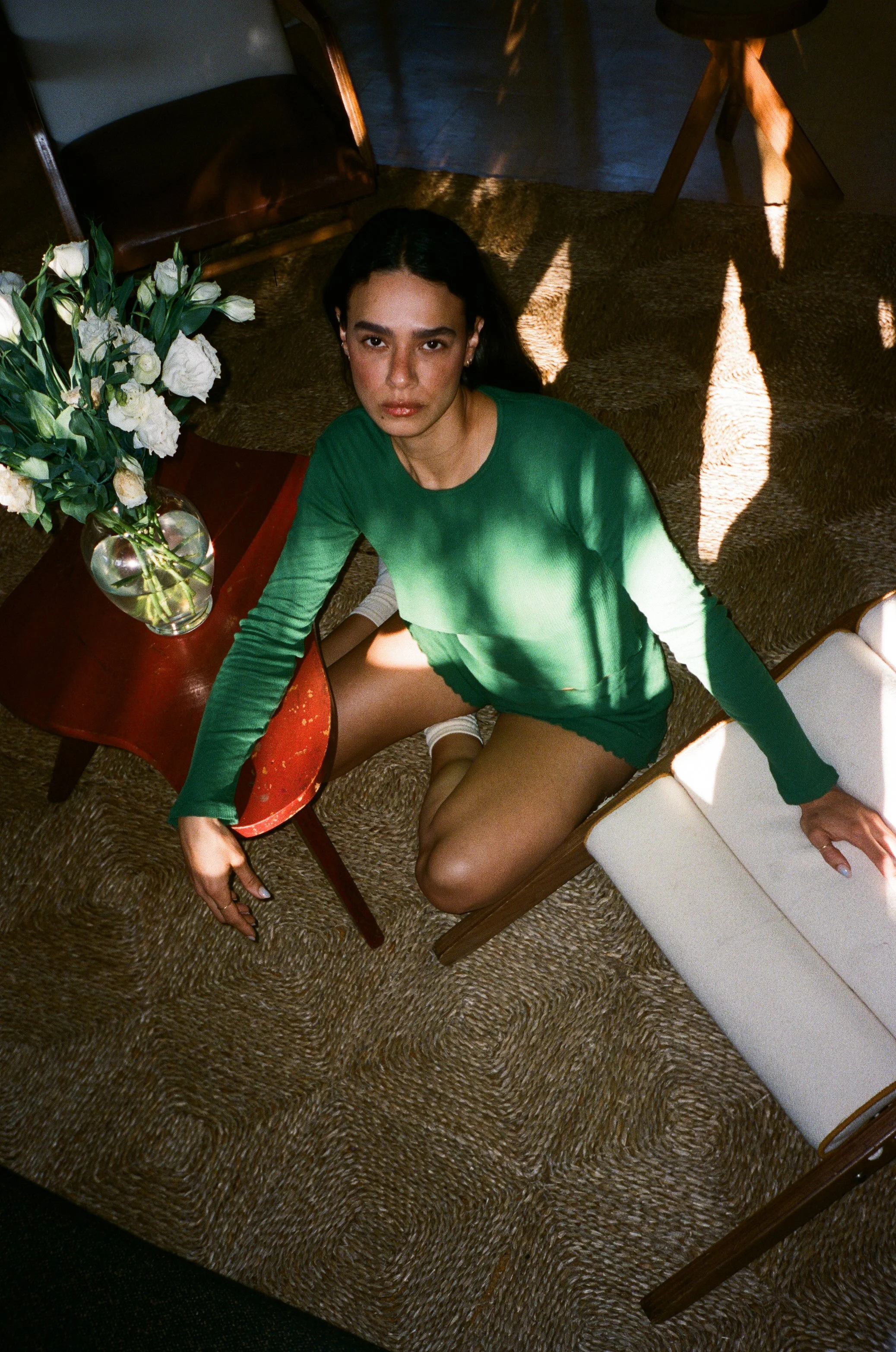 A woman with dark hair in a green long-sleeve top and shorts sitting on the floor next to a small red table with a vase of white flowers, in a room with patterned carpet and sunlight casting shadows.