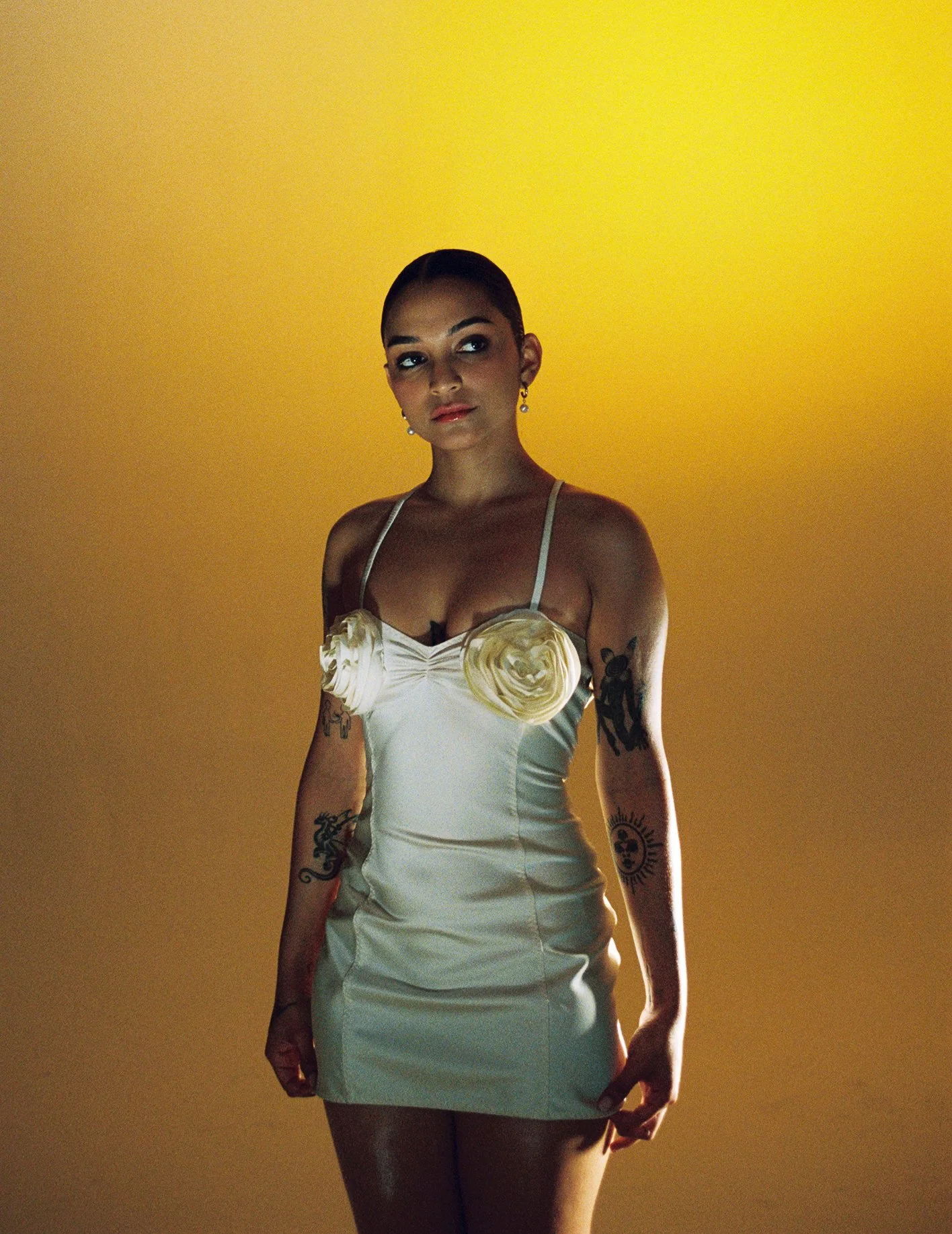 A woman with tattoos and a short dress standing against a yellow background.