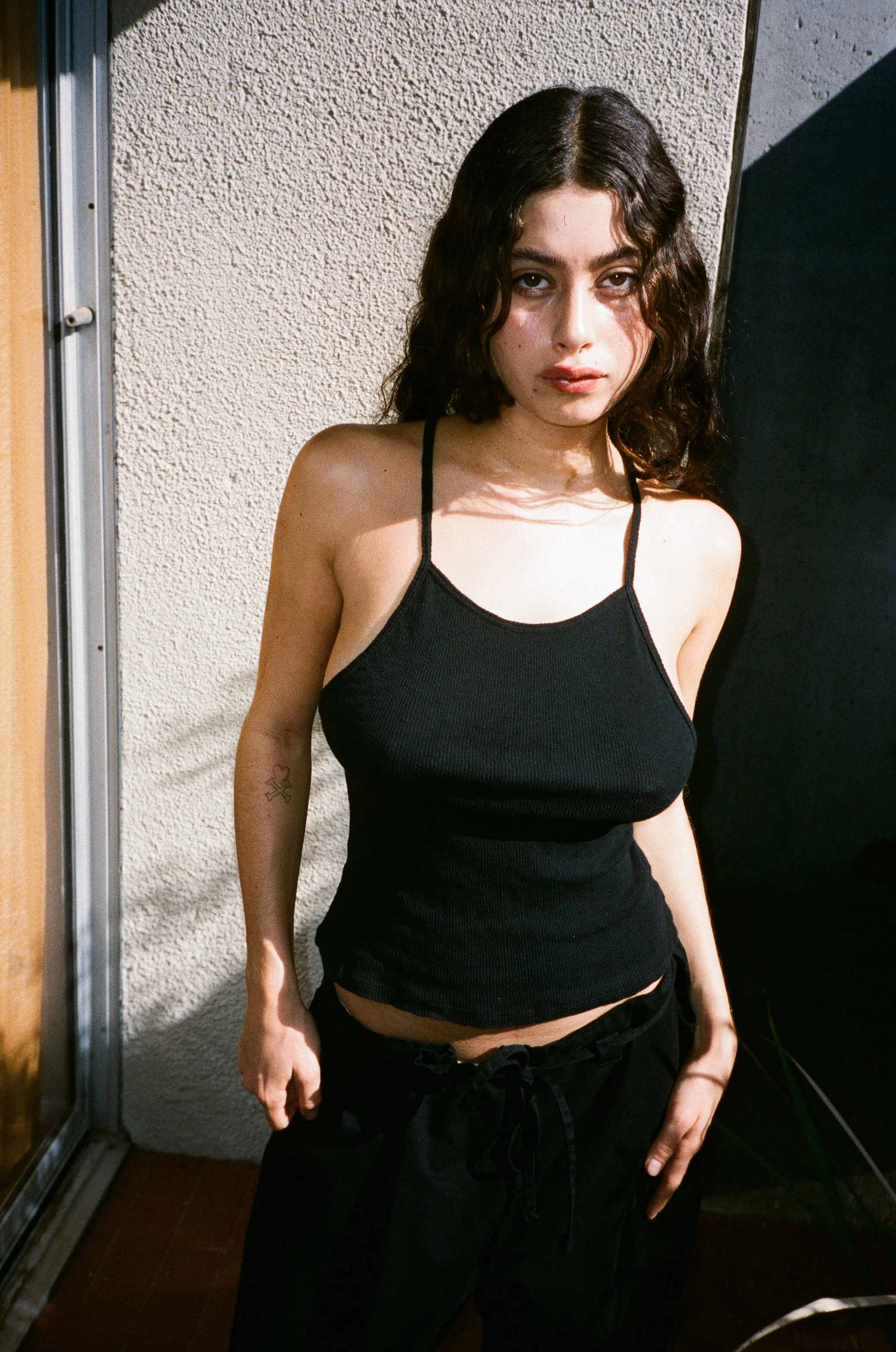 Young woman with dark, wavy hair wearing a black tank top and black pants standing outdoors near a textured wall and window.