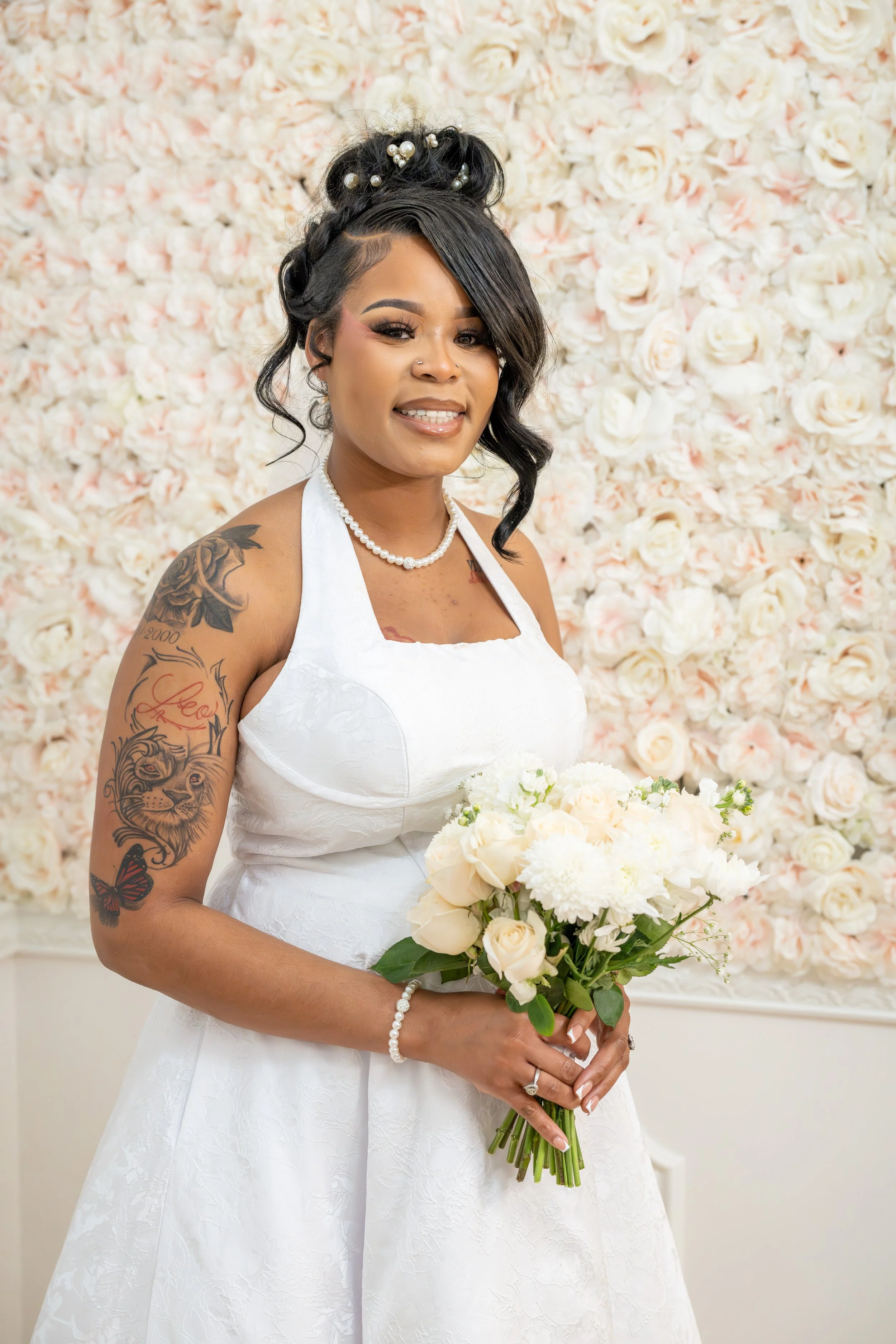 A woman in a white wedding dress holding a bouquet of white flowers, standing in front of a wall of white roses with pink accents.