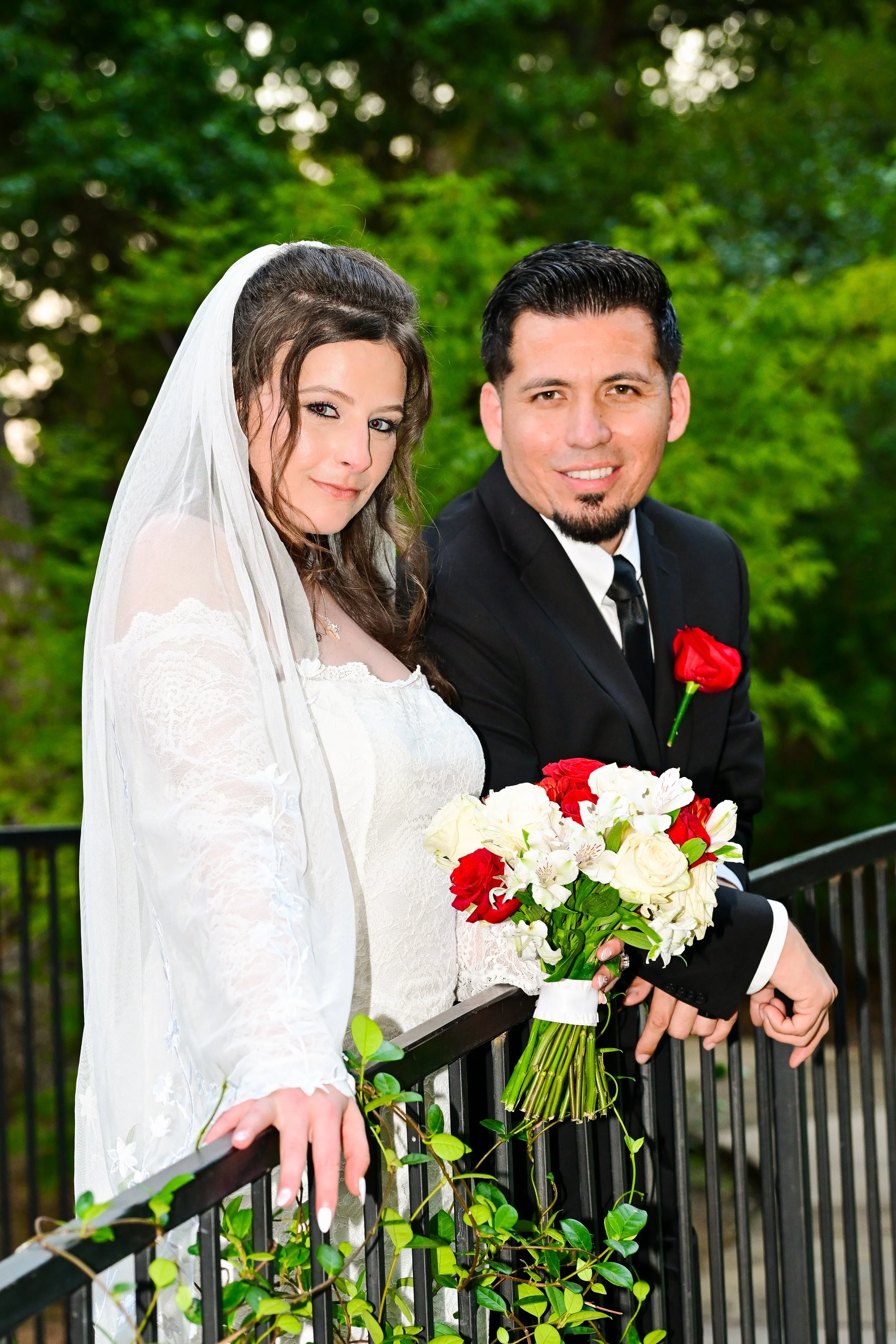 A bride in a white wedding dress and veil holding a bouquet of red and white flowers, standing next to a groom in a black suit with a red boutonniere, outdoors with green trees in the background.