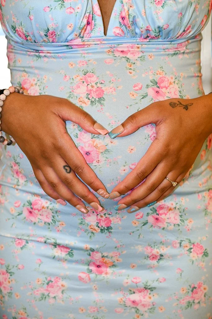 A woman wearing a light blue dress with pink floral patterns forming a heart shape with her hands over her belly.