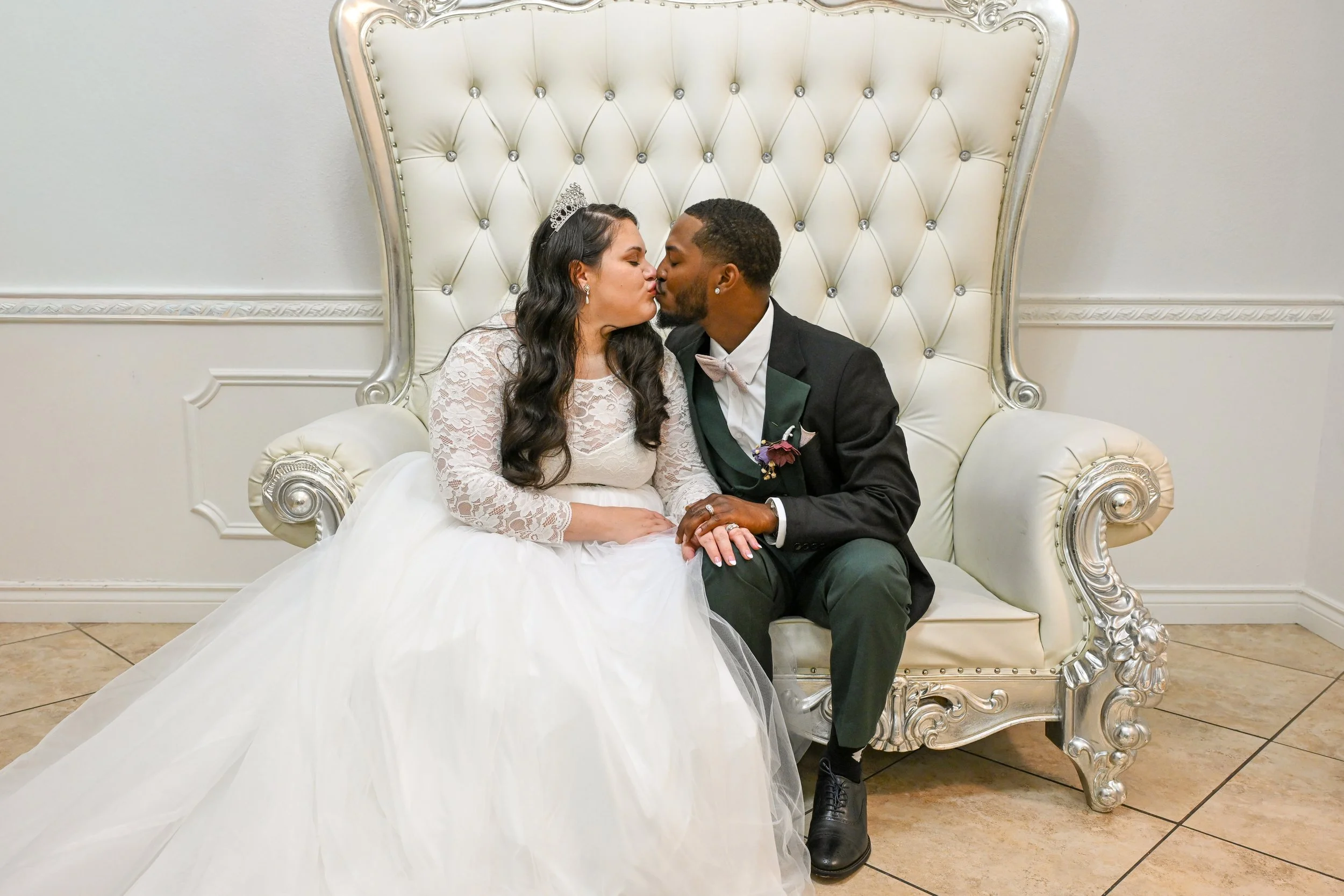 A bride and groom sitting on a white ornate couch, sharing a kiss, at their wedding reception. The bride is wearing a white lace wedding gown with long sleeves and a tiara, and the groom is in a black tuxedo with a pink bow tie and boutonniere.