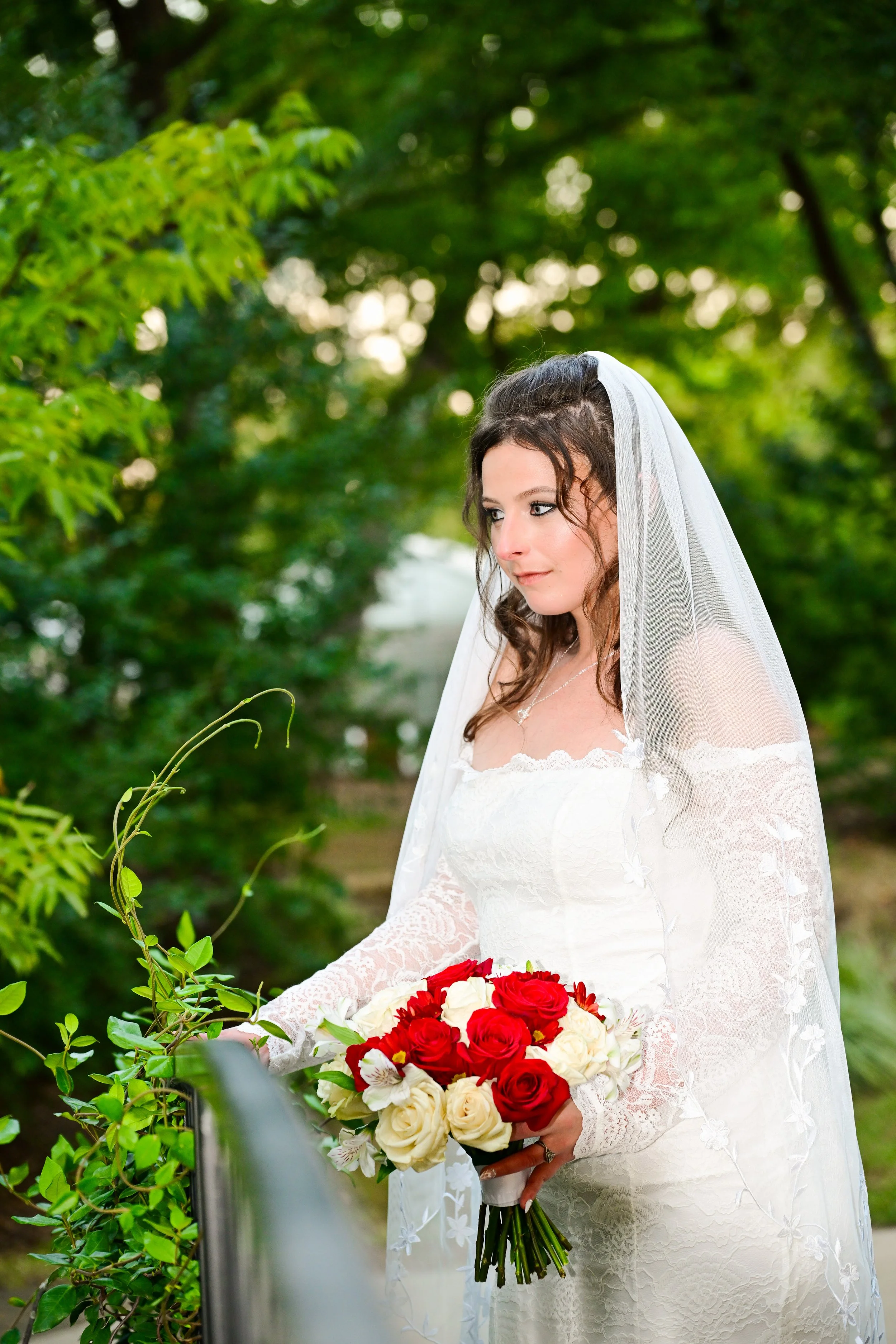 Bride in white wedding dress with lace sleeves and veil holding a bouquet of red and white roses, outdoors among green trees.