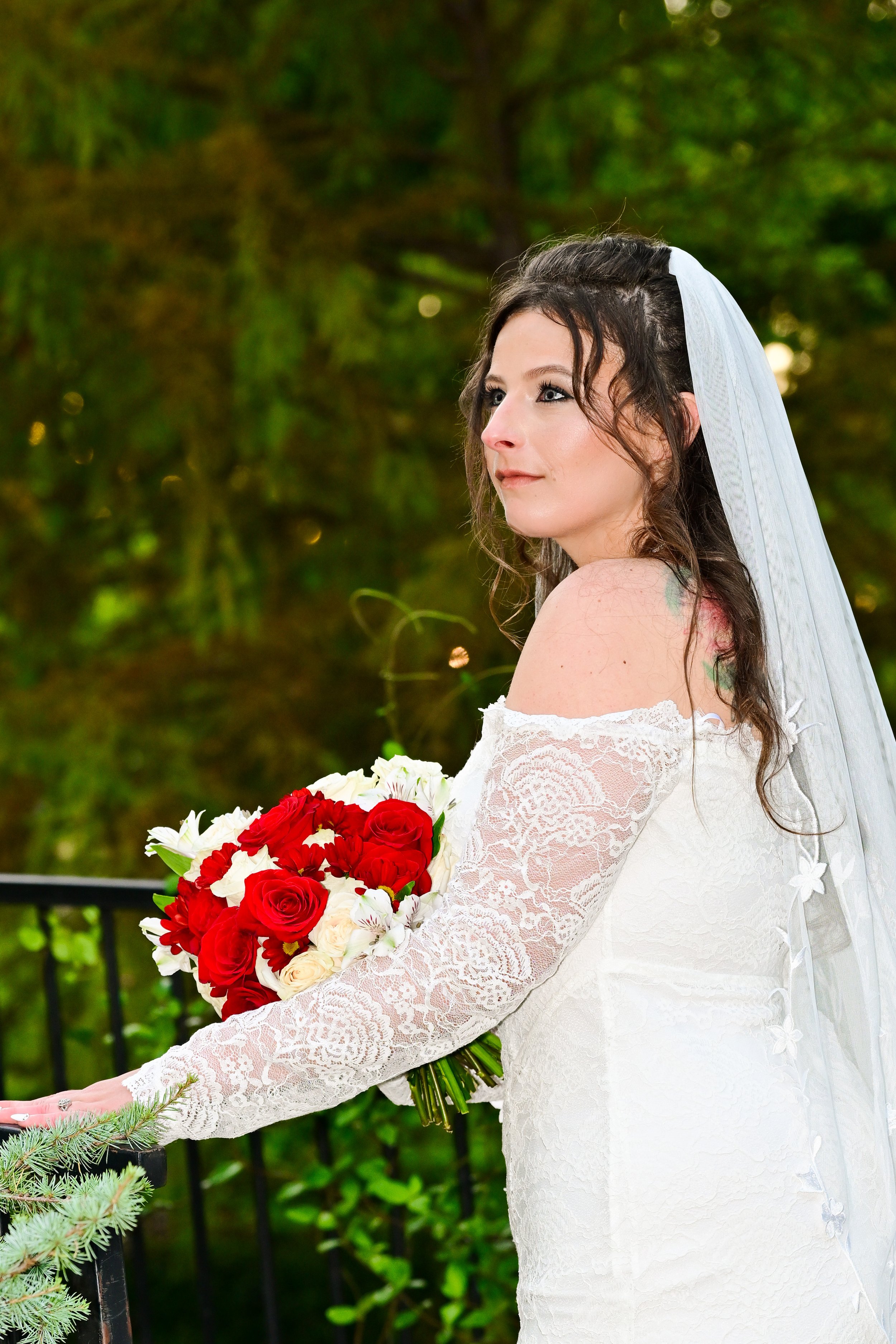 A bride with long brown hair and a white lace off-shoulder wedding dress, holding a bouquet of red and white roses, standing outdoors with green trees in the background.