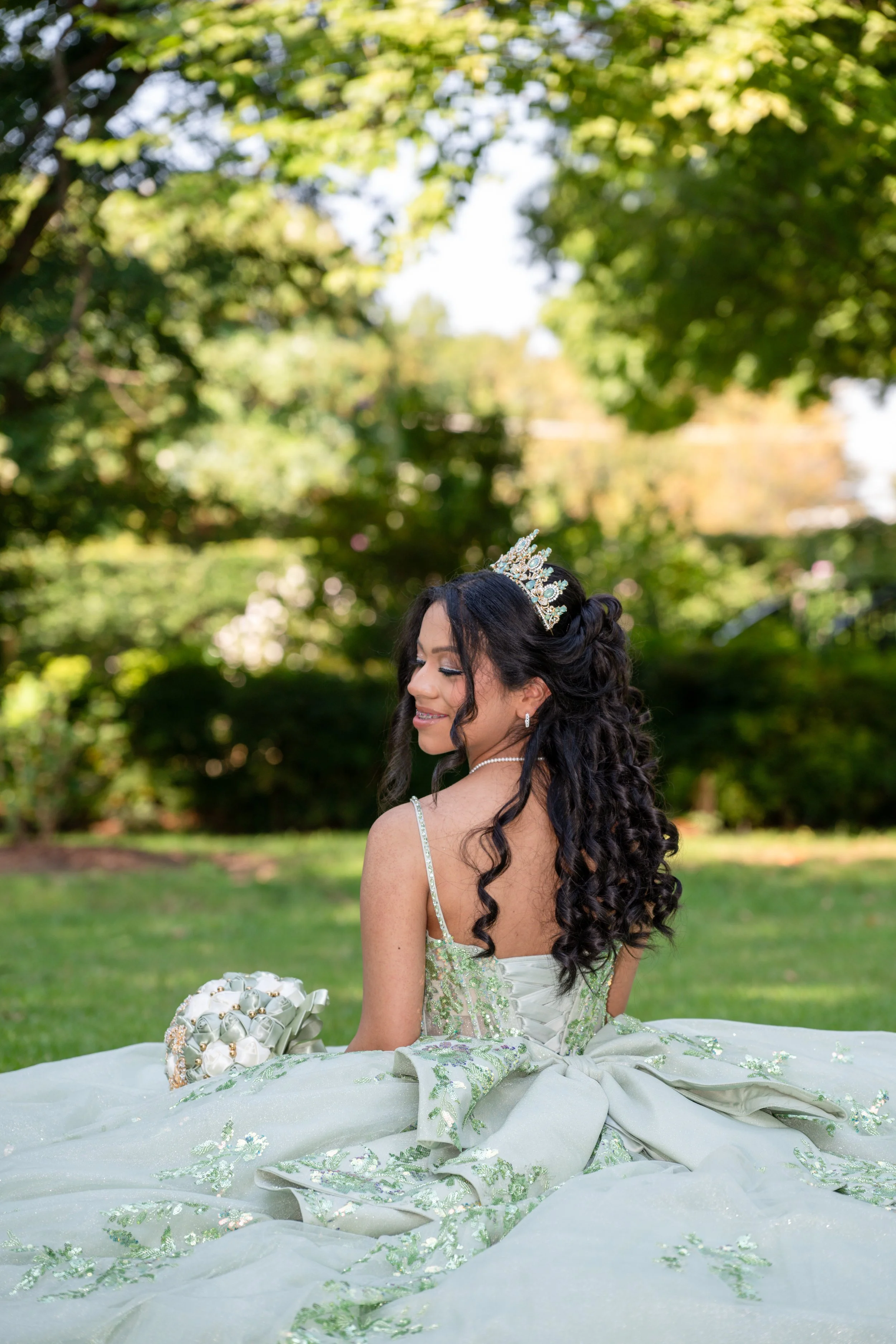 A woman in a wedding dress with a tiara, sitting outdoors on green grass with trees in the background.