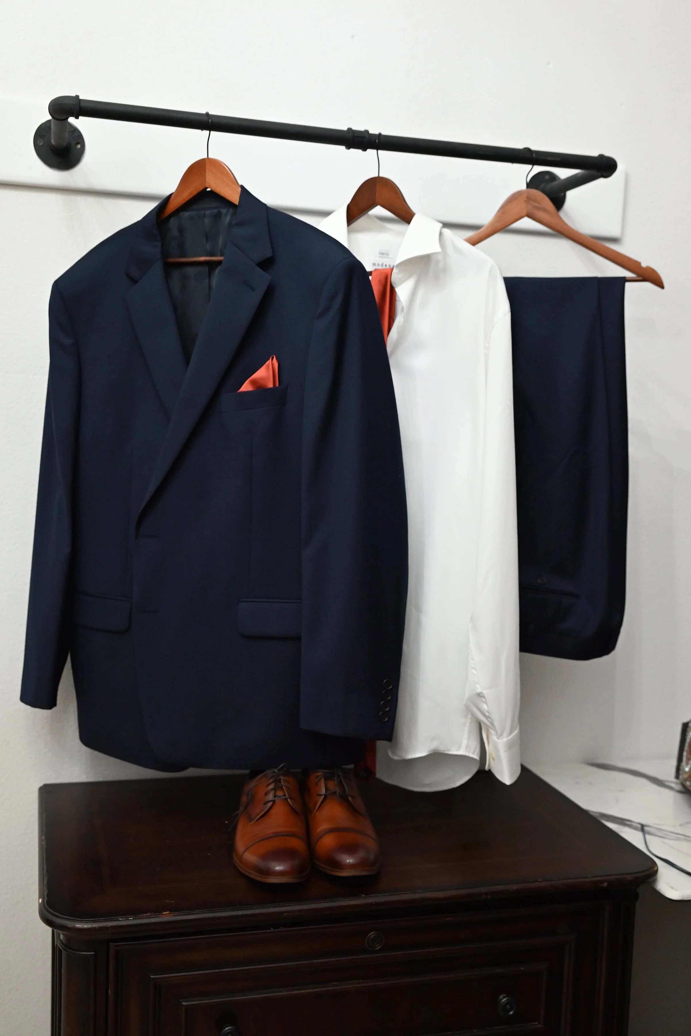 Navy suit with a peach pocket square, white dress shirt, pair of navy dress pants, and brown dress shoes on a dark wooden surface, all hanging on wooden and black hangers against a white wall.