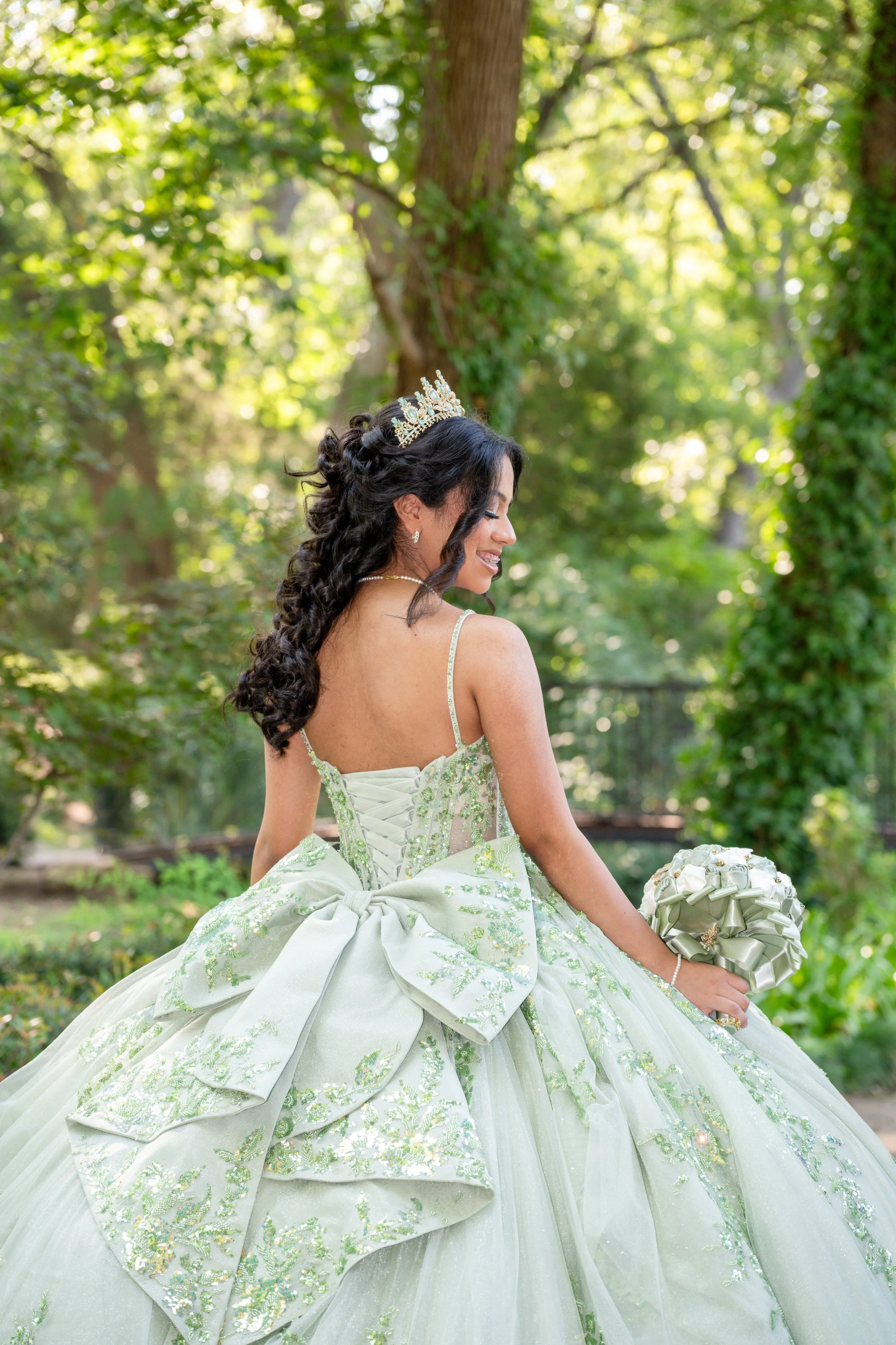 A woman in a light green wedding gown with a large bow on the back, holding a bouquet, sitting outdoors in a lush, green, wooded area, wearing a tiara and jewelry.