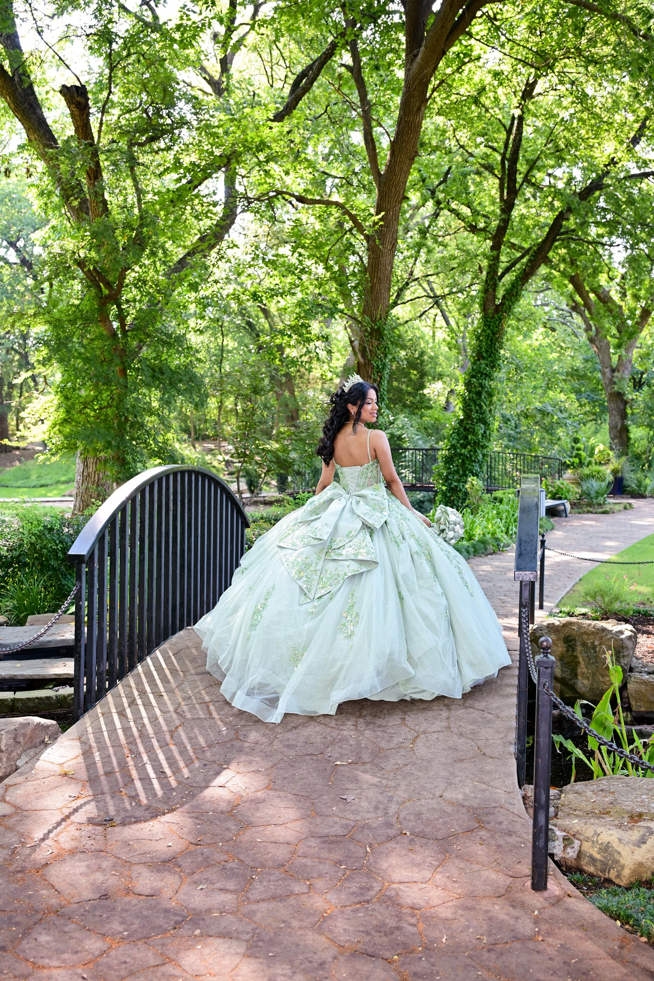 A young woman in a light green, elaborate ball gown with a large bow back, standing on a stone bridge in a lush green park surrounded by tall trees and greenery.