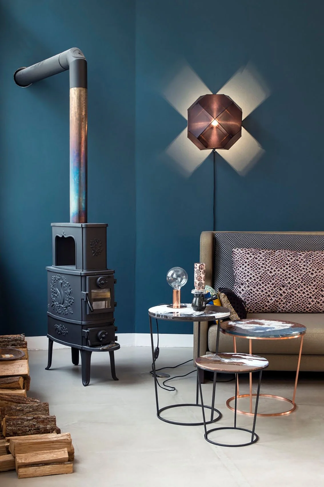 Interior of a stylish room featuring a dark teal wall, a modern wall sconce with geometric copper design, a beige sofa with patterned pillows, a small black and white round marble side table with decorative items, a vintage black stove with a pipe, and a stack of firewood in the foreground.