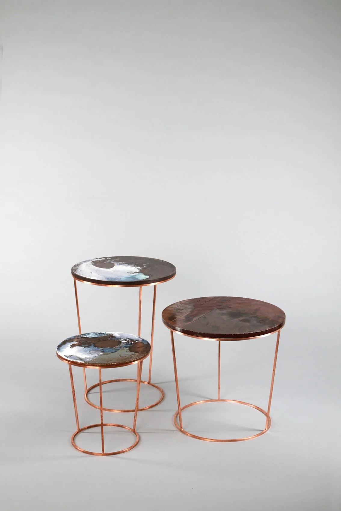 Set of three round tables with copper-colored metal frames, two tables featuring dark marbled tops, one with a painted outer surface, arranged against a plain light gray background.