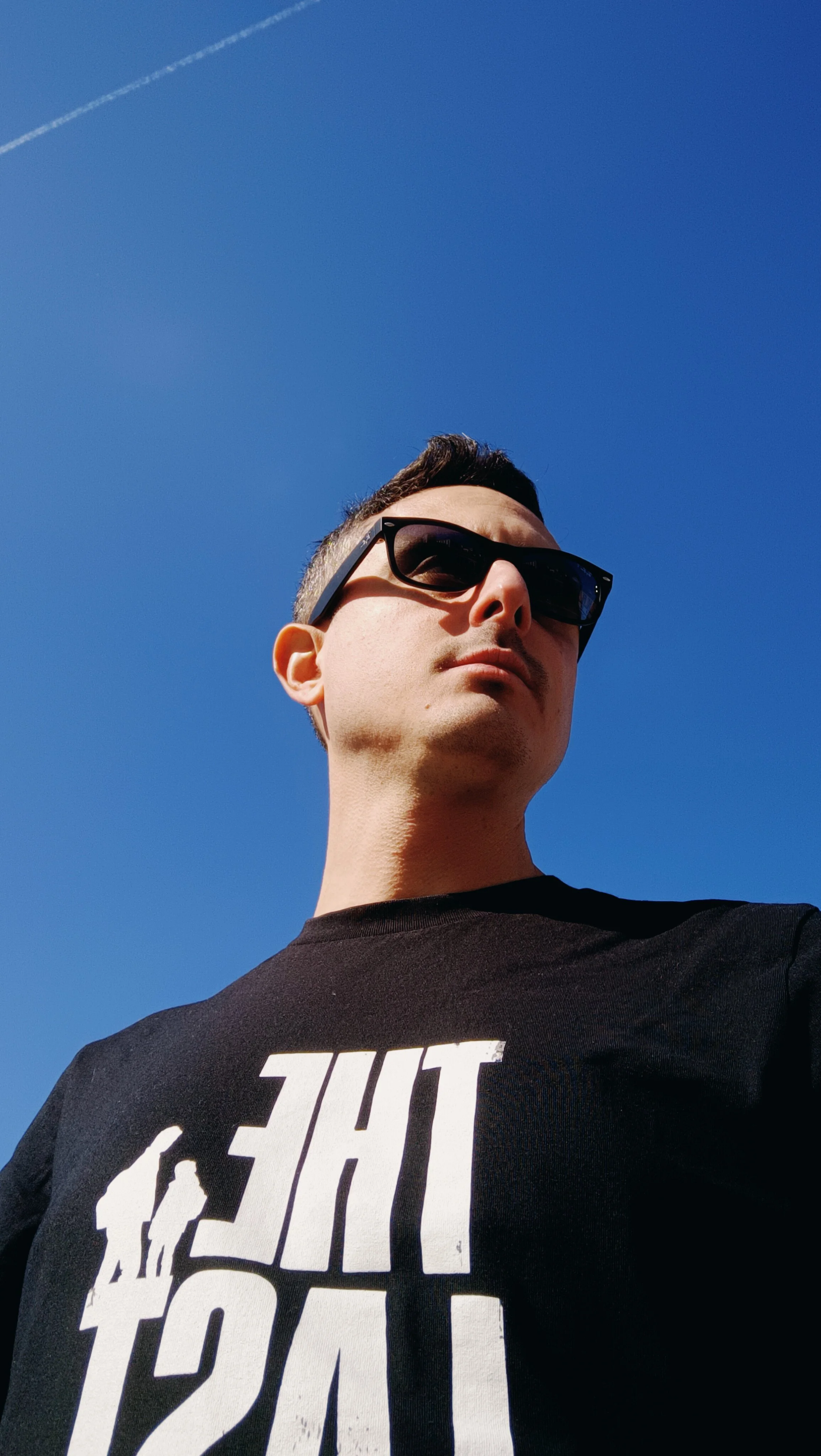Un uomo indossa occhiali da sole e una maglietta nera con scritte bianche, sotto un cielo blu chiaro.