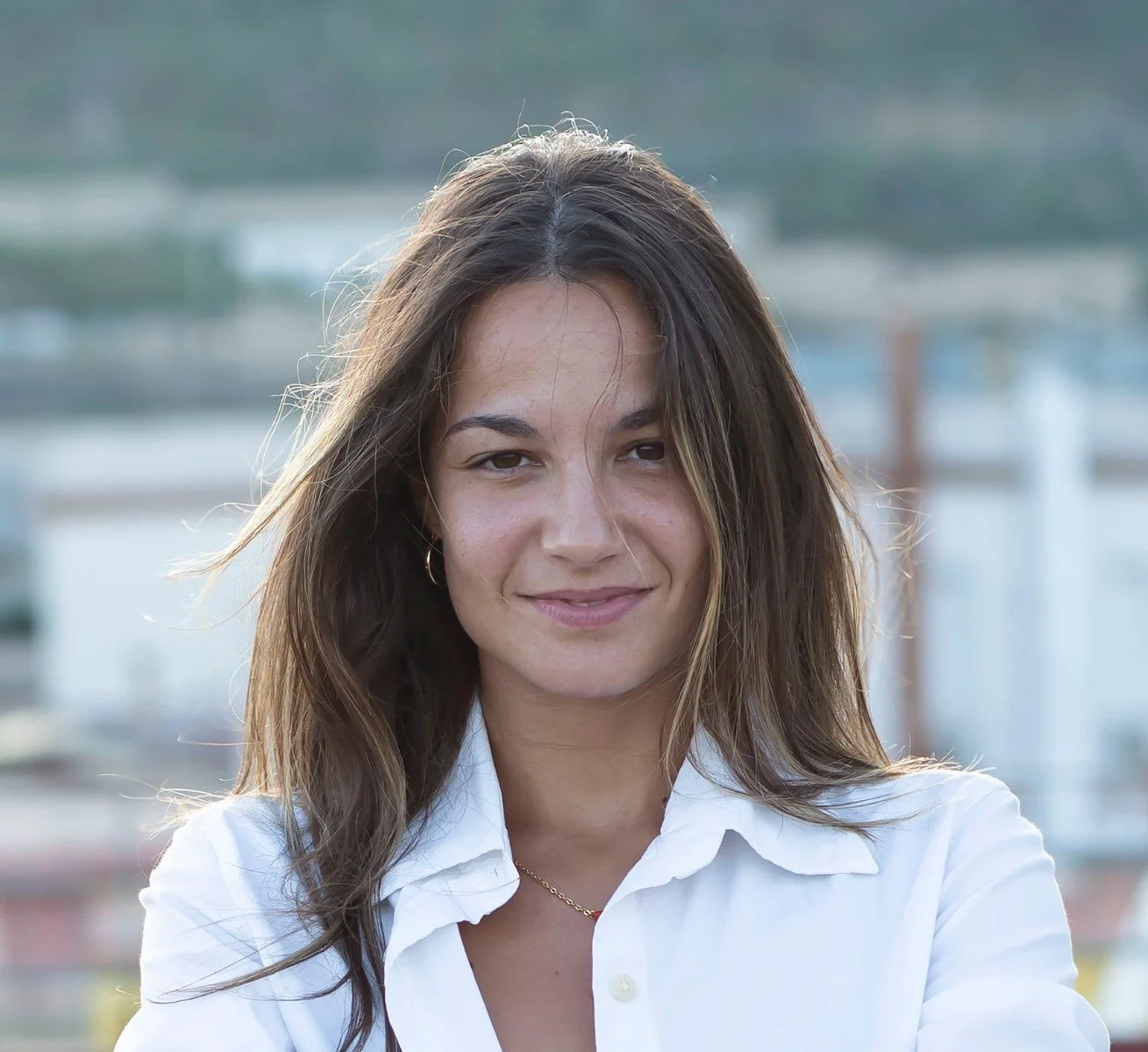 Ragazza sorridente con capelli castani, indossa una camicia bianca e orecchini d'oro, sfondo all'aperto.