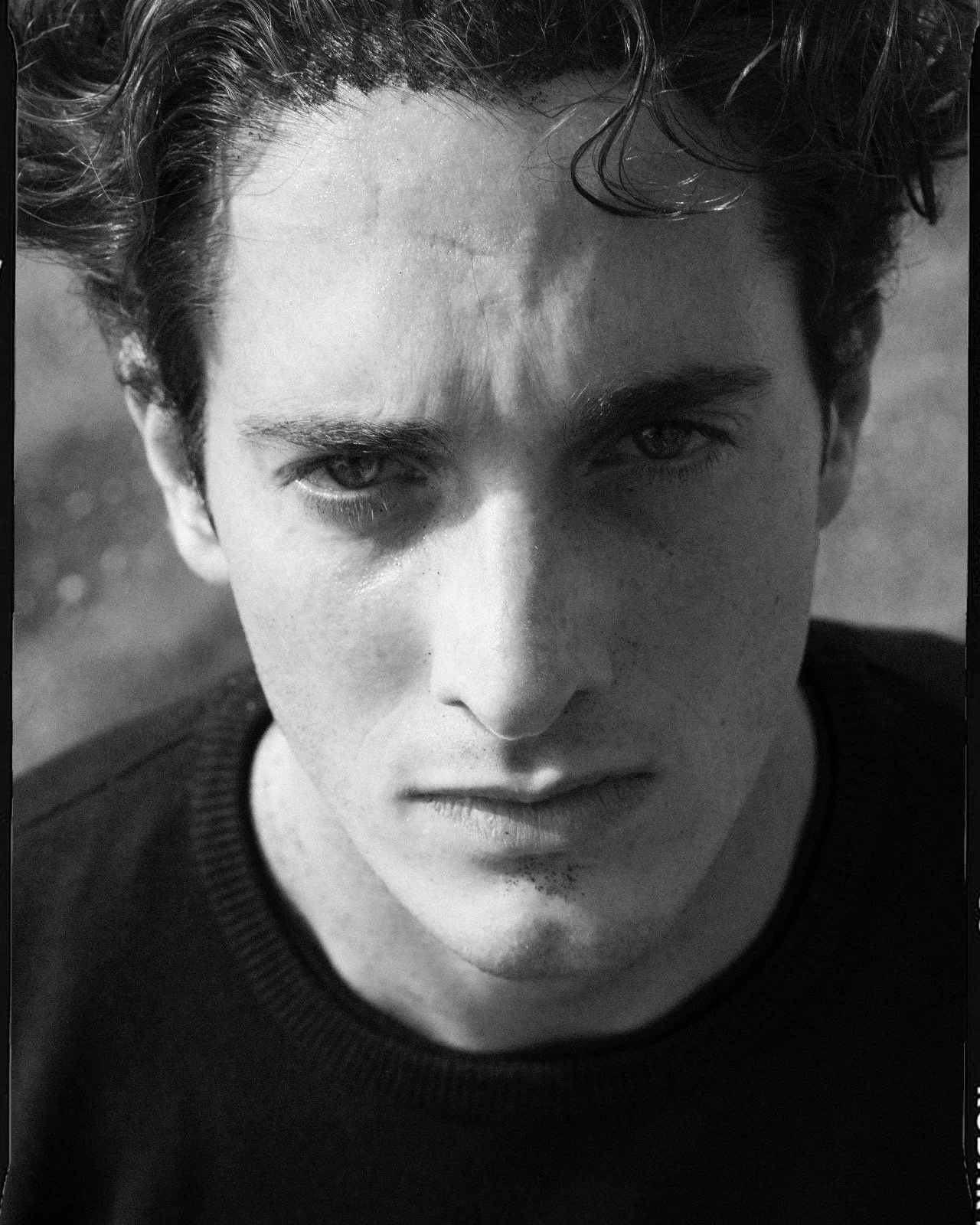 Ritratto in bianco e nero di un giovane uomo con capelli mossi, sguardo intenso e labbra chiuse, che indossa una maglietta scura.