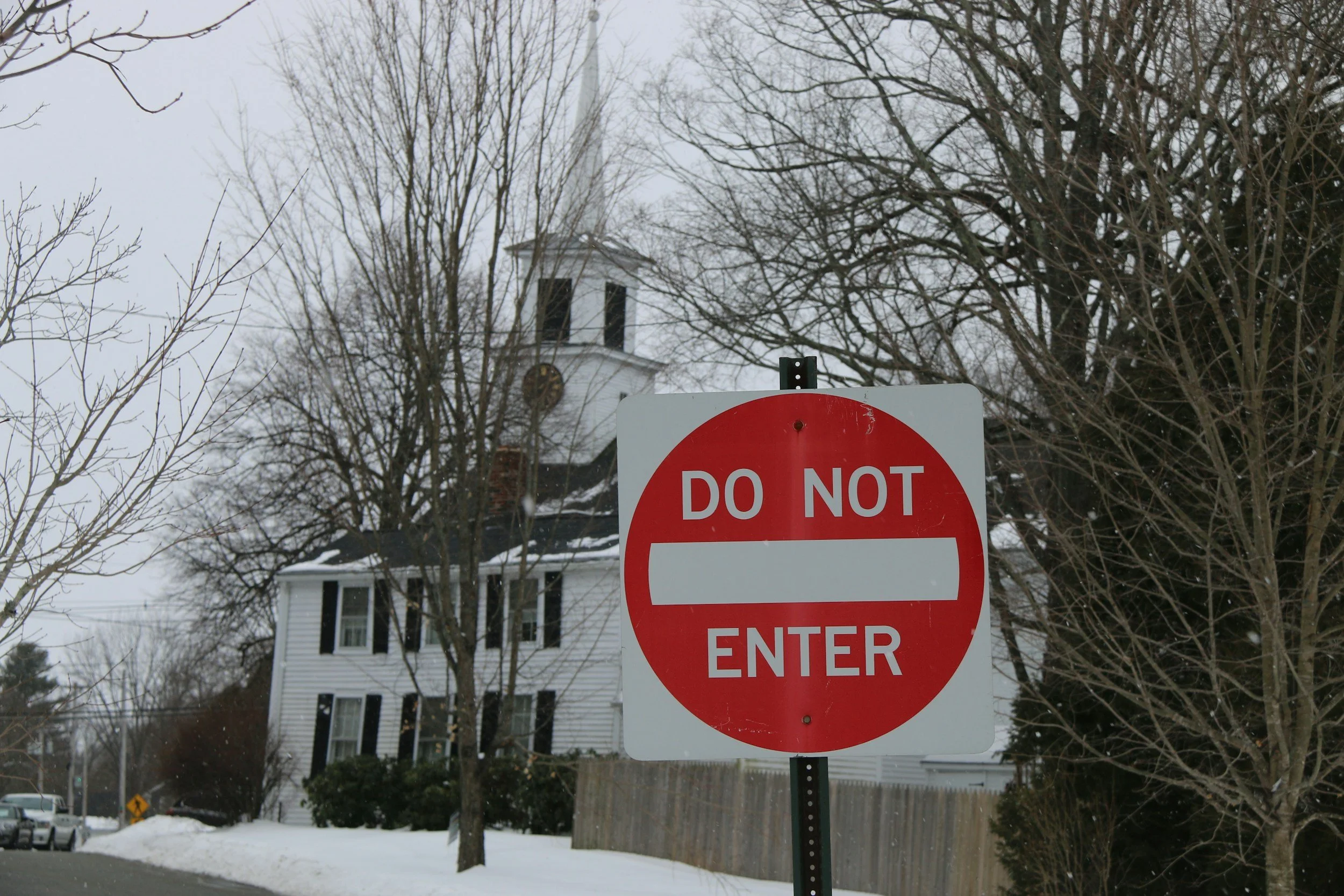 A red and white 'Do Not Enter' traffic sign in a snow-covered residential neighborhood with leafless trees, houses, and parked cars in the background.