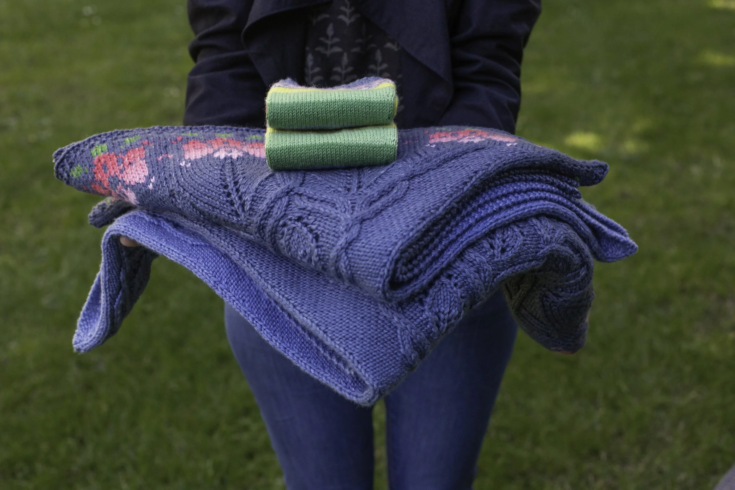 Andrea is holding a pile of blue knitted fabric with cables and stranded colourwork. On top of the pile is what looks like a neatly folded pair of socks.