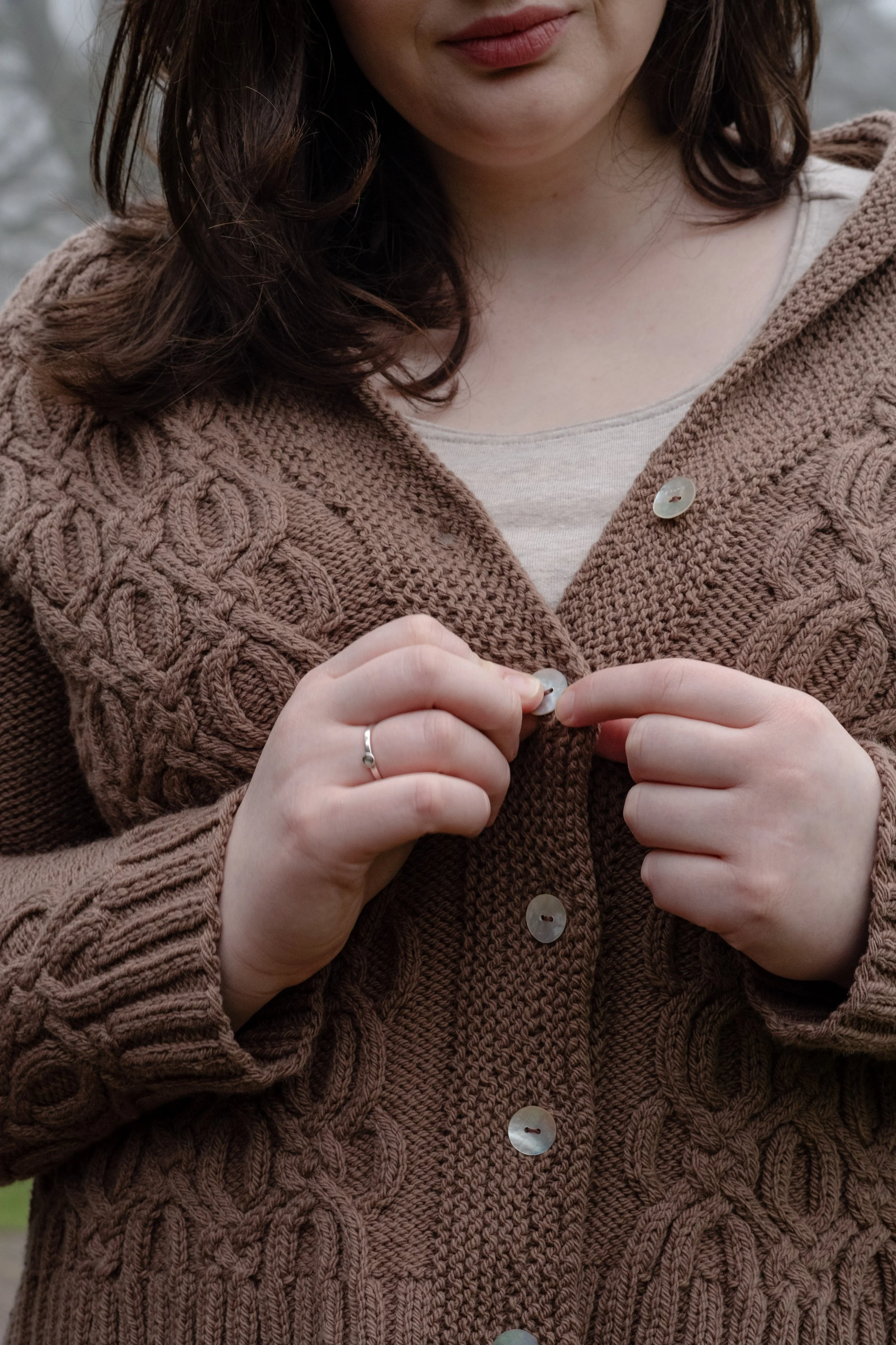 Closeup of Kelsey buttoning a richly-cabled brown sweater.