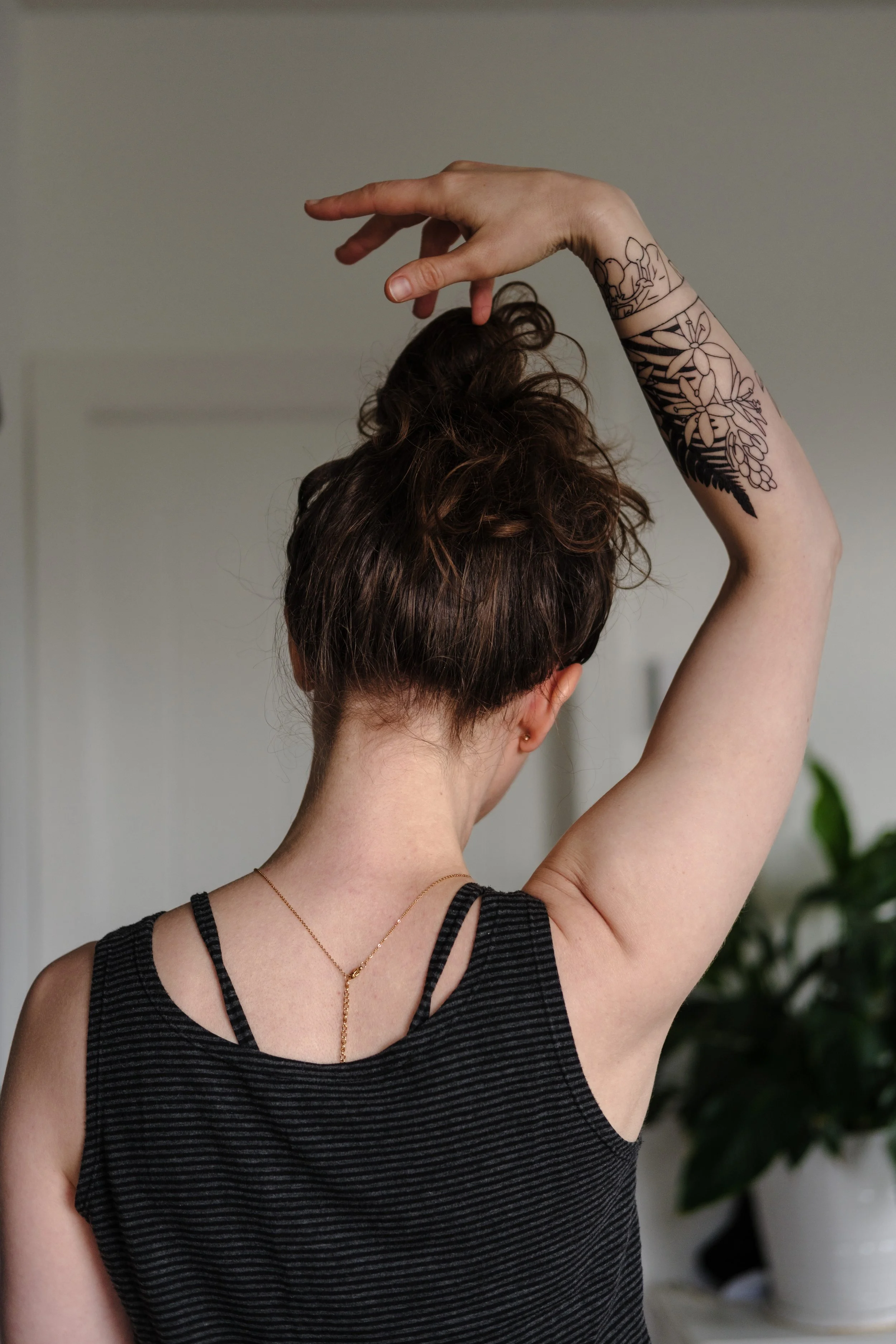 Andrea from the back. Her right arm is raised above her head displaying a tattoo around her forearm with camas and salal flowers and a dark, shaded fern. An empty band separates the tattoo into two parts.